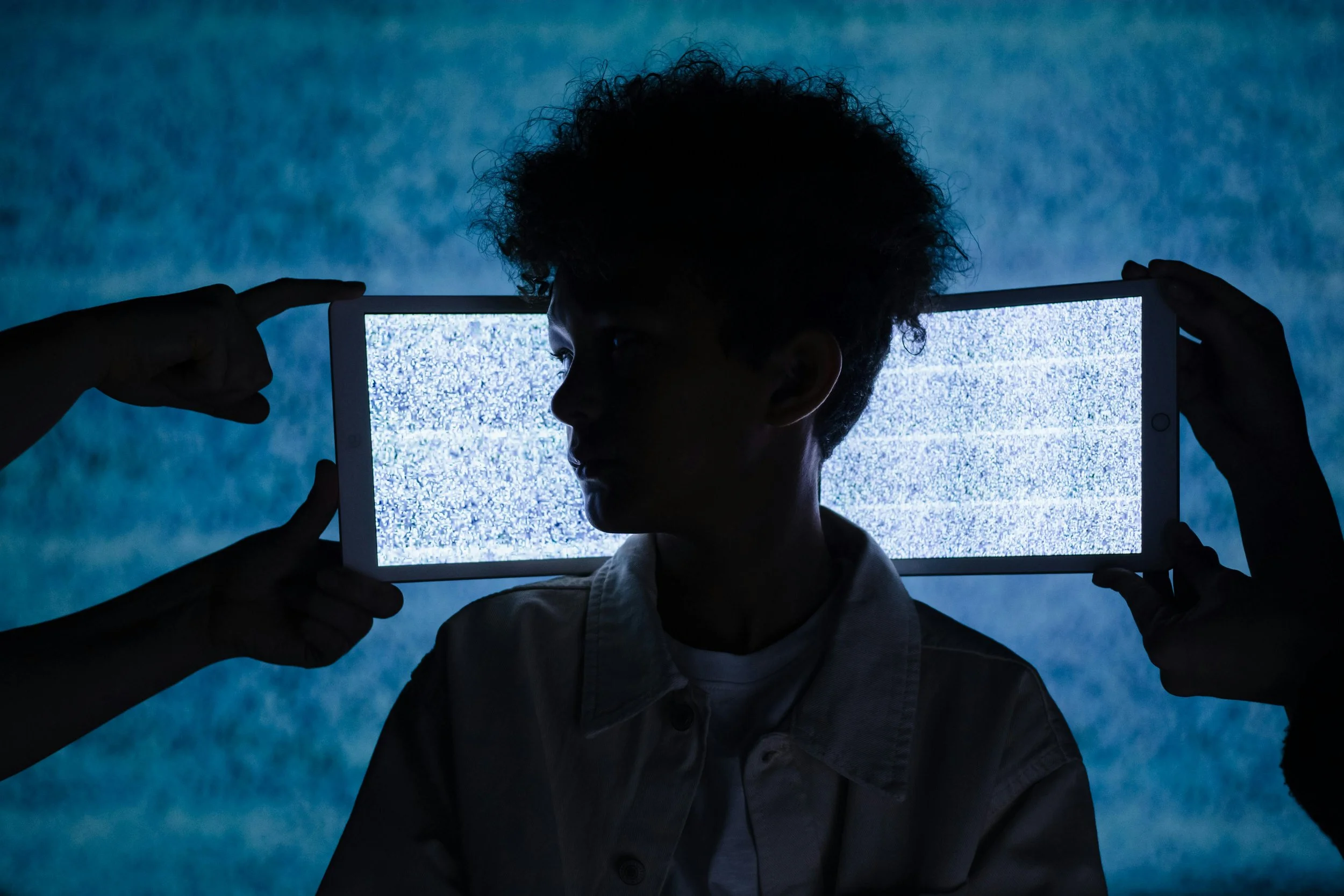 A silhouetted woman stands against a blue background while a bright tablet screen frames their face, with two hands pointing toward their head, representing digital pressure and online harassment.