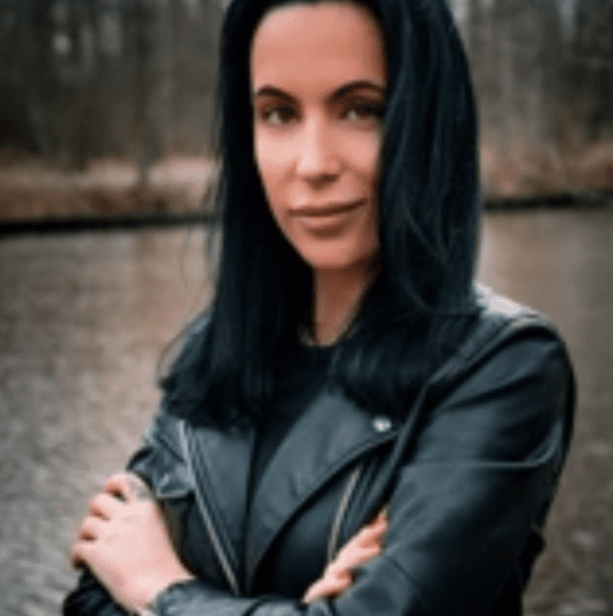 Portrait of a woman with dark hair wearing a black jacket, standing outdoors by water with trees in the background.