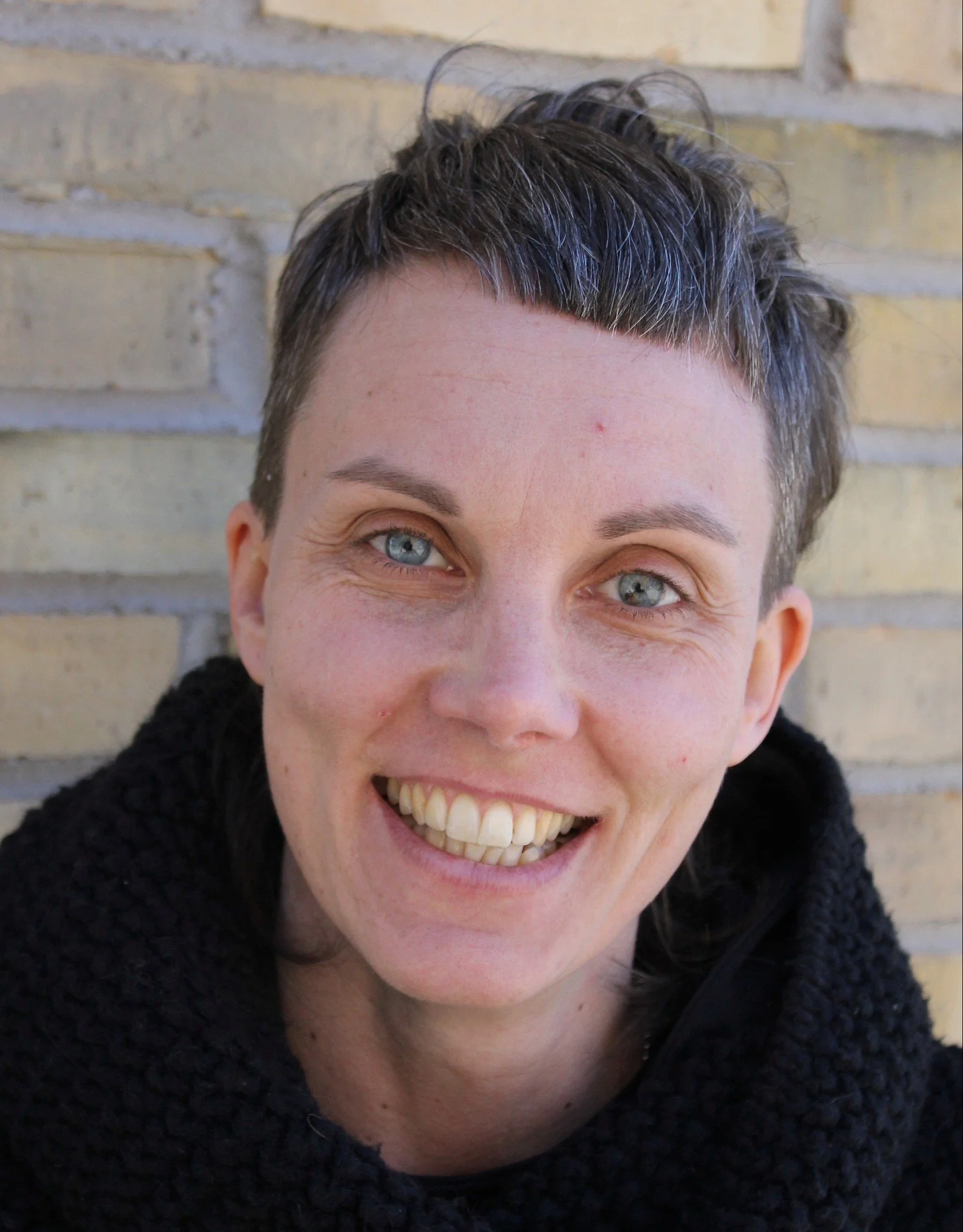 Close-up outdoor portrait of a woman with short dark hair and blue eyes, smiling broadly, wearing a dark coat, with a light brick wall in the background.
