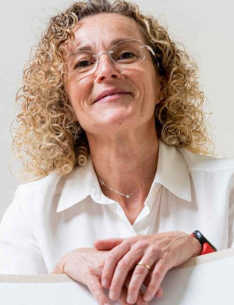 Portrait of a woman with shoulder-length curly blonde hair and glasses, wearing a light blouse, leaning forward with her hands resting on a surface and smiling gently toward the camera.
