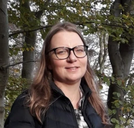 Outdoor portrait of a woman with long light brown hair and glasses, wearing a dark jacket, standing among trees and foliage and looking toward the camera.