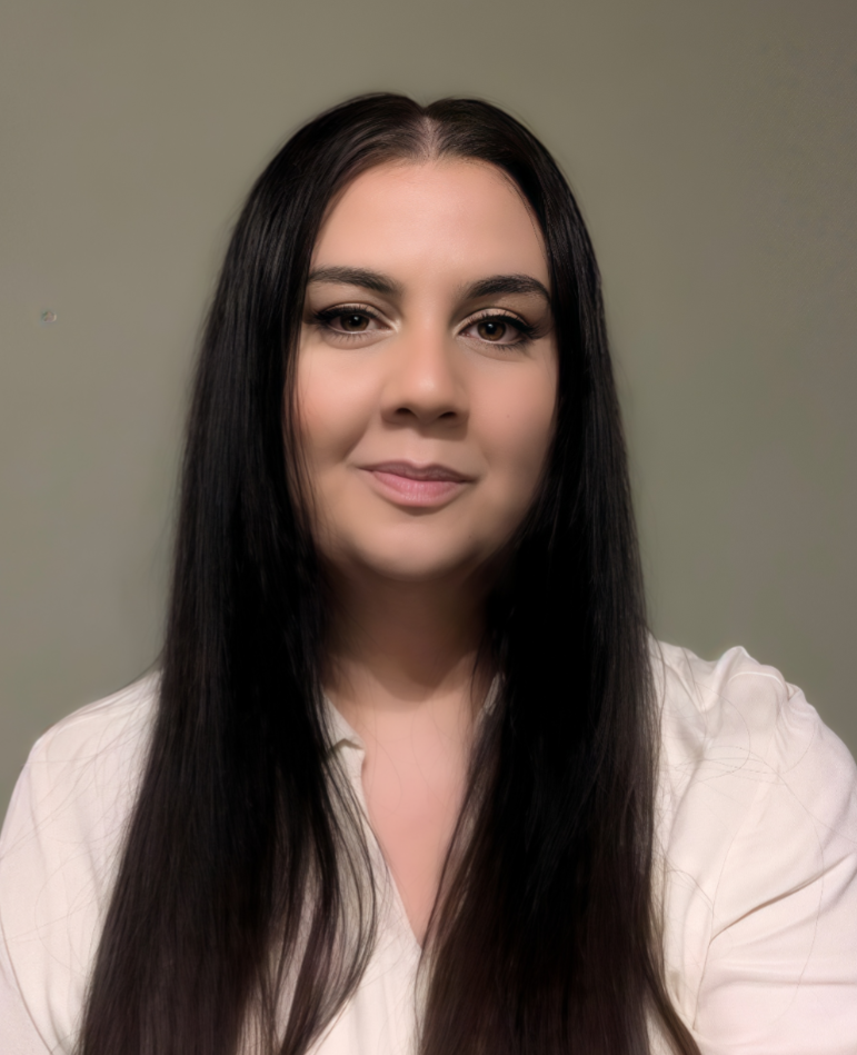 Studio-style portrait of a woman with long dark hair, wearing a light blouse, facing the camera with a neutral expression against a plain background.