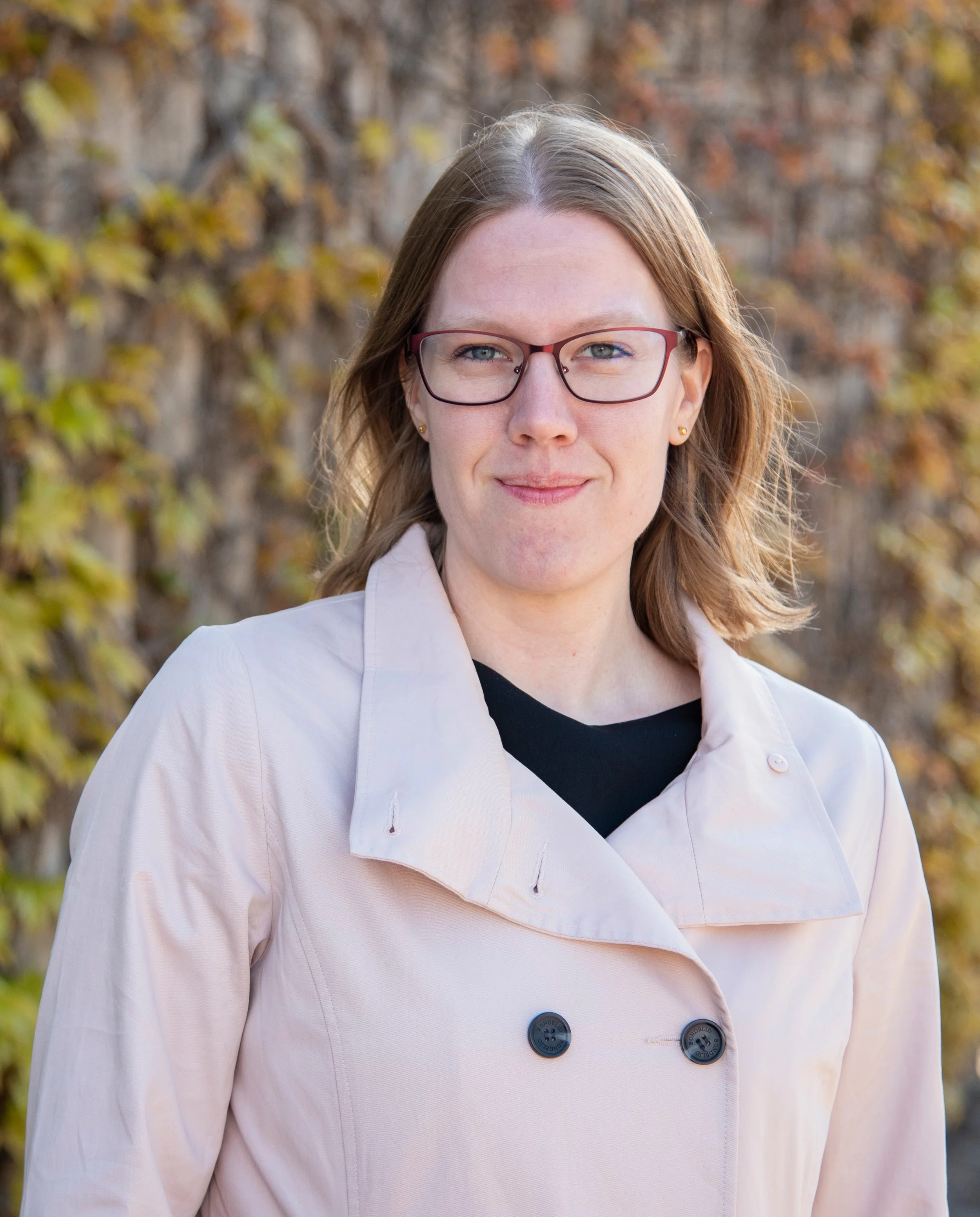 Outdoor portrait of a woman with shoulder-length light brown hair and glasses, wearing a pale coat over a dark top, standing in front of a wall covered in autumn leaves.