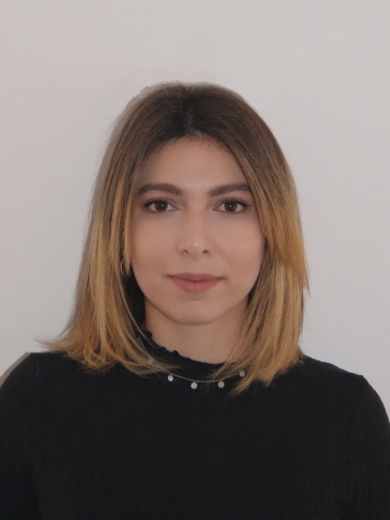 Portrait of a woman with shoulder-length light brown hair, wearing a black top and necklace, facing the camera with a neutral expression against a plain light background.