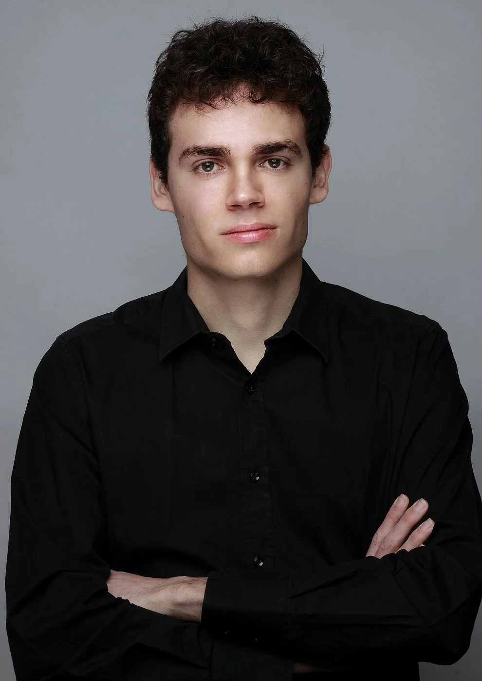 Studio portrait of a person with short dark curly hair wearing a black buttoned shirt, standing with arms crossed and looking directly at the camera against a plain grey background.