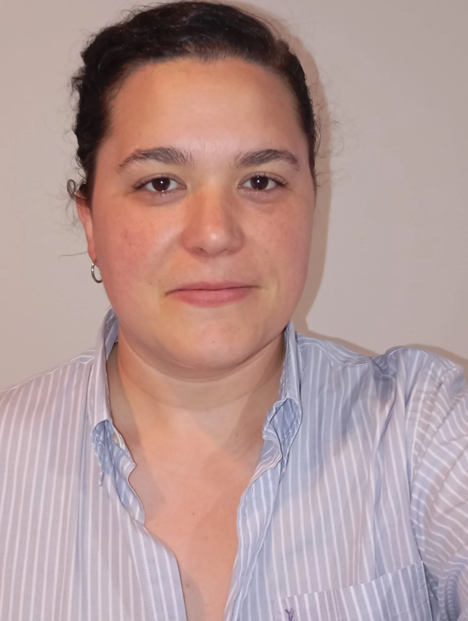 Close-up portrait of a woman with dark hair pulled back, wearing a light blue striped shirt and small hoop earrings, facing the camera with a neutral expression against a plain light-coloured background.