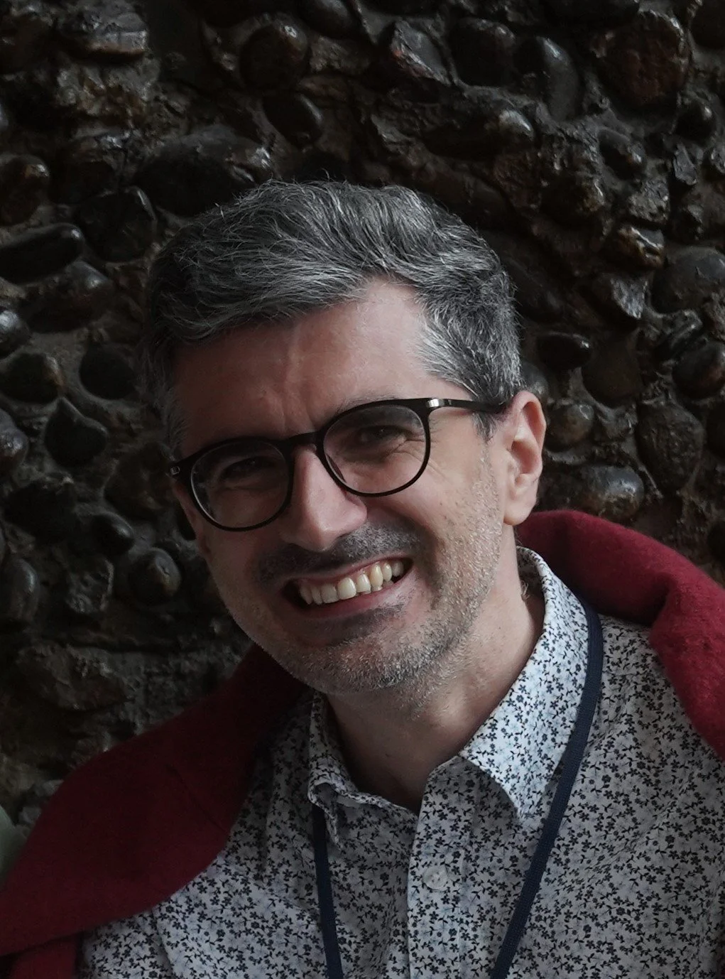 Outdoor portrait of a man with short greying hair and glasses, smiling, wearing a patterned shirt and jacket, standing against a textured stone wall.