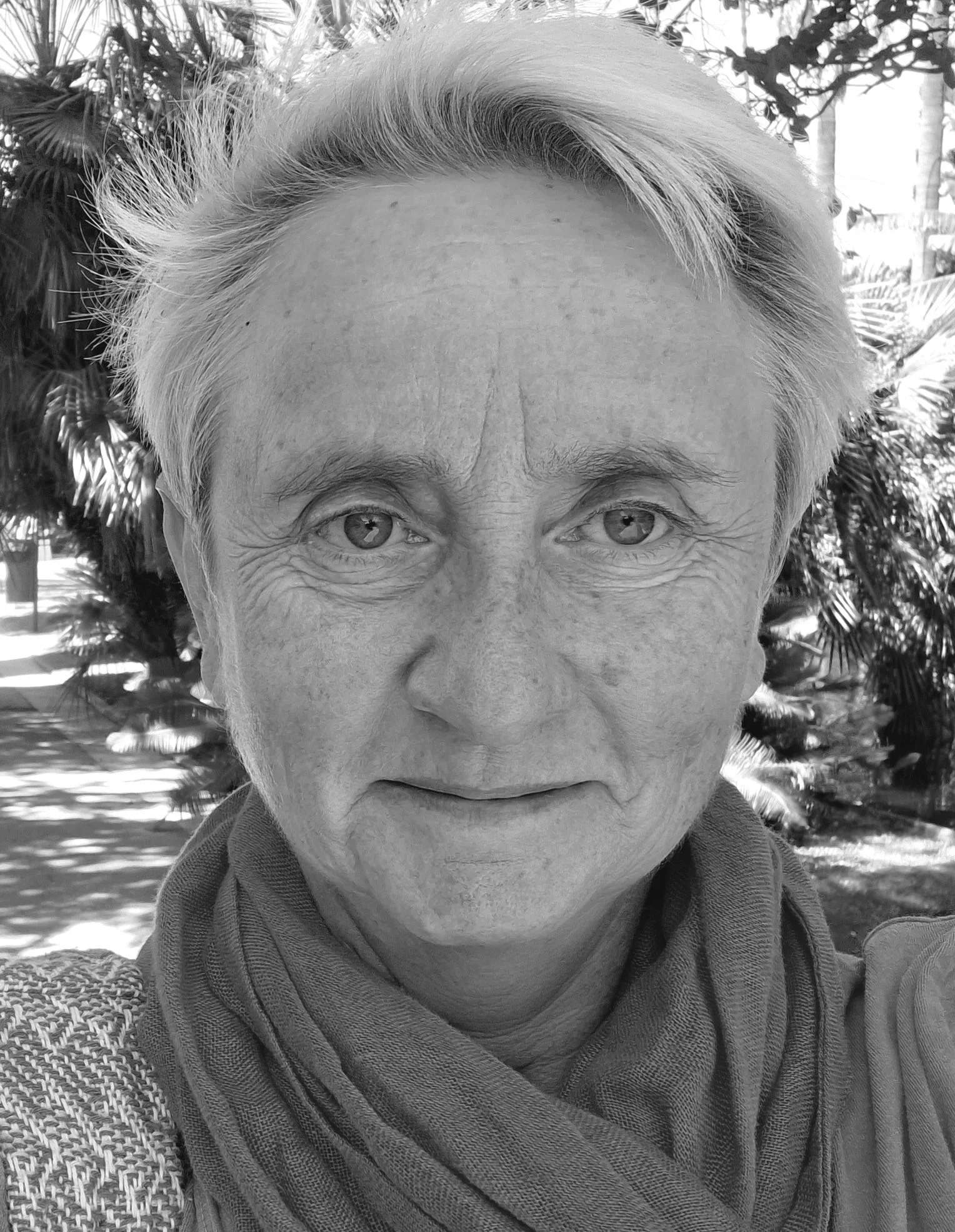 Black-and-white outdoor portrait of an older woman with short light hair, wearing a scarf and textured jacket, looking directly at the camera with trees and foliage in the background.  If you want, I can: