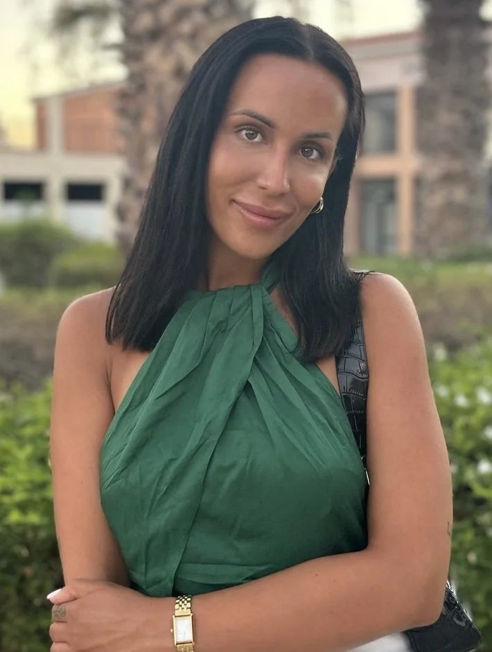 Portrait of a woman wearing a green top, standing outdoors in front of greenery and palm trees.