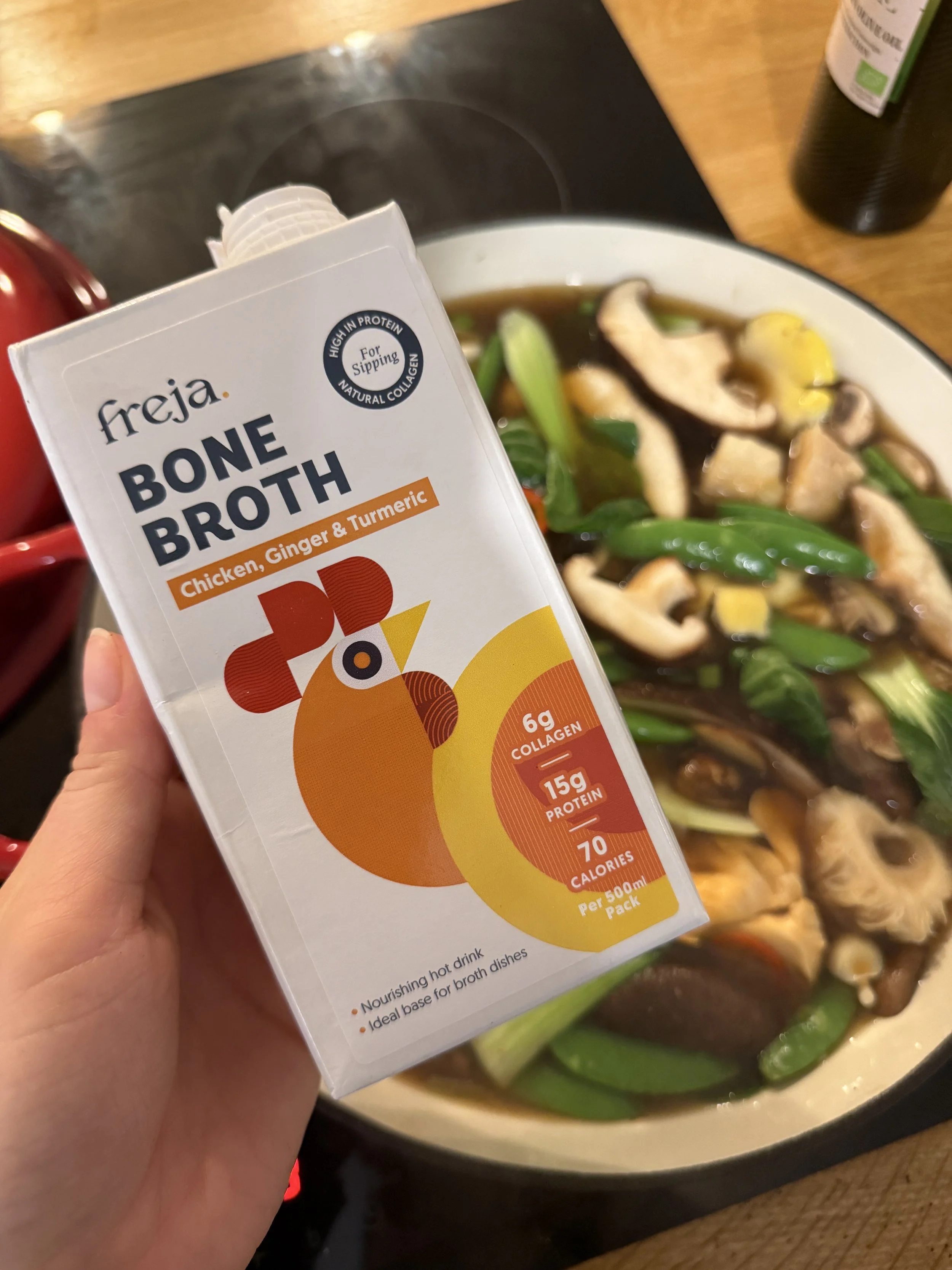A person holding a carton of Freja bone broth with chicken, ginger, and turmeric in front of a pot of soup with vegetables and mushrooms.