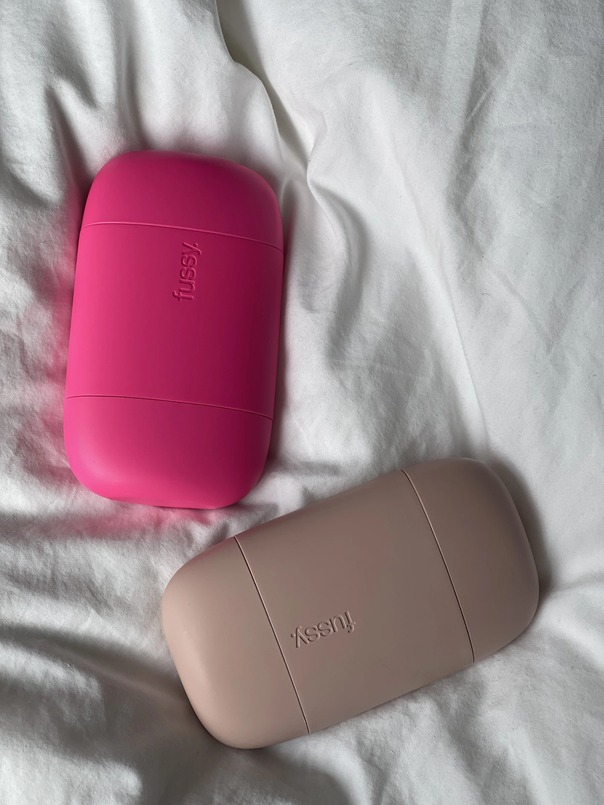 Close-up of two Hello Sunday products on white fabric, one pink and one beige, both with sleek, rounded design.