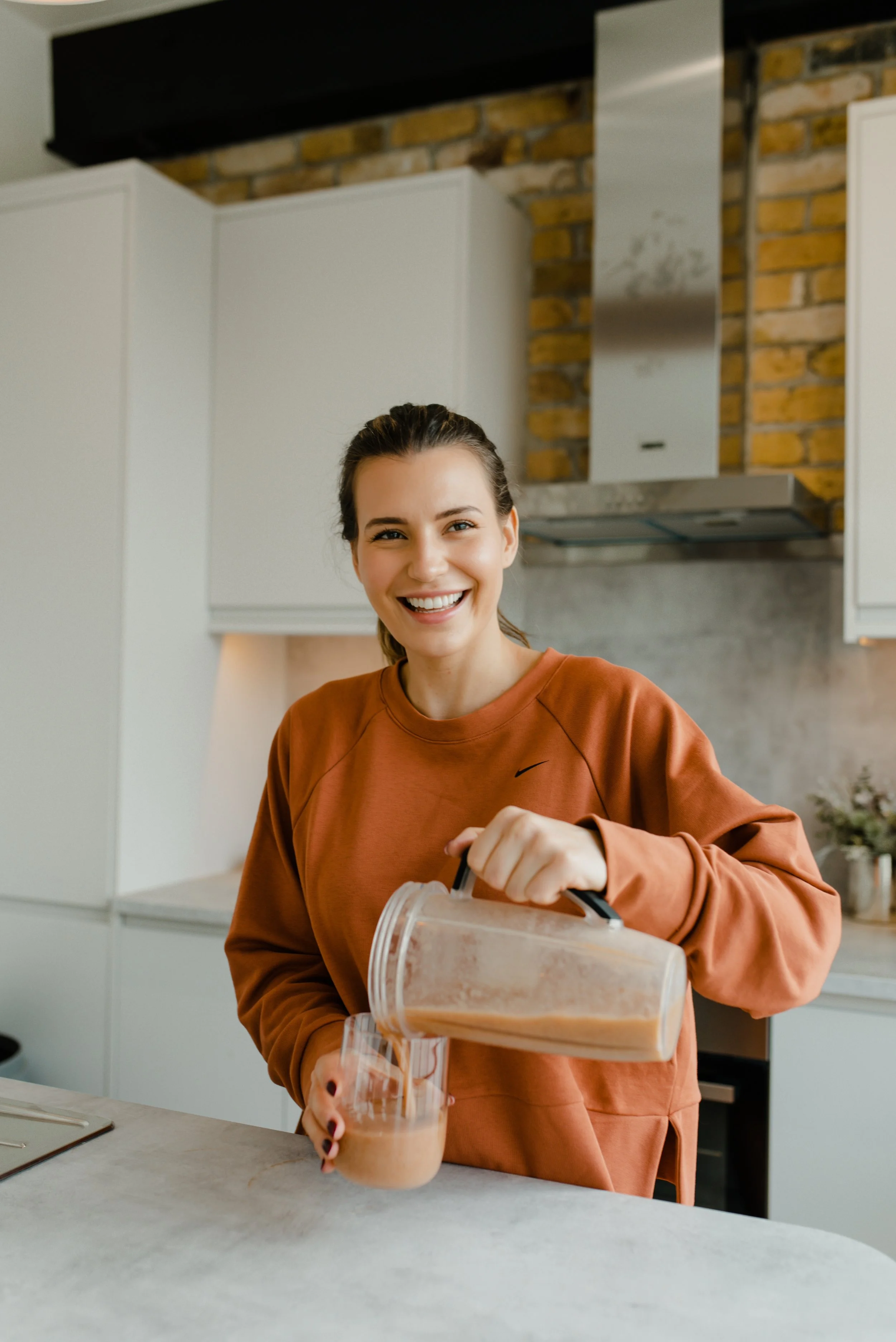 A young woman with brown hair and a brown sweatshirt smiling as she pours iced coffee into a glass in a modern kitchen.