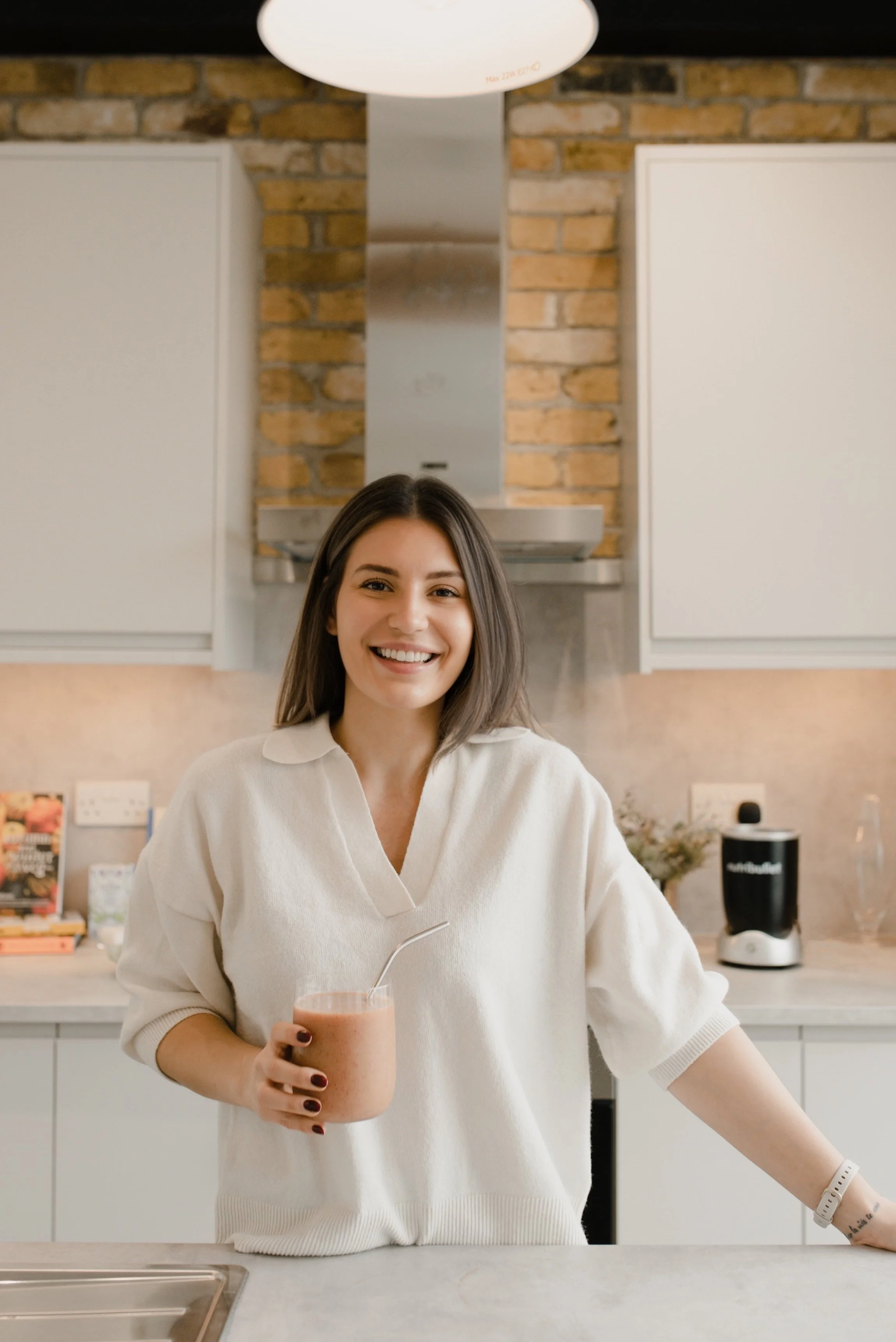 A woman with shoulder-length brown hair smiling and holding a pink smoothie with a straw in a kitchen. She is wearing a white shirt and has a tattoo on her left forearm. The background shows kitchen cabinets, a brick wall, and a blender.