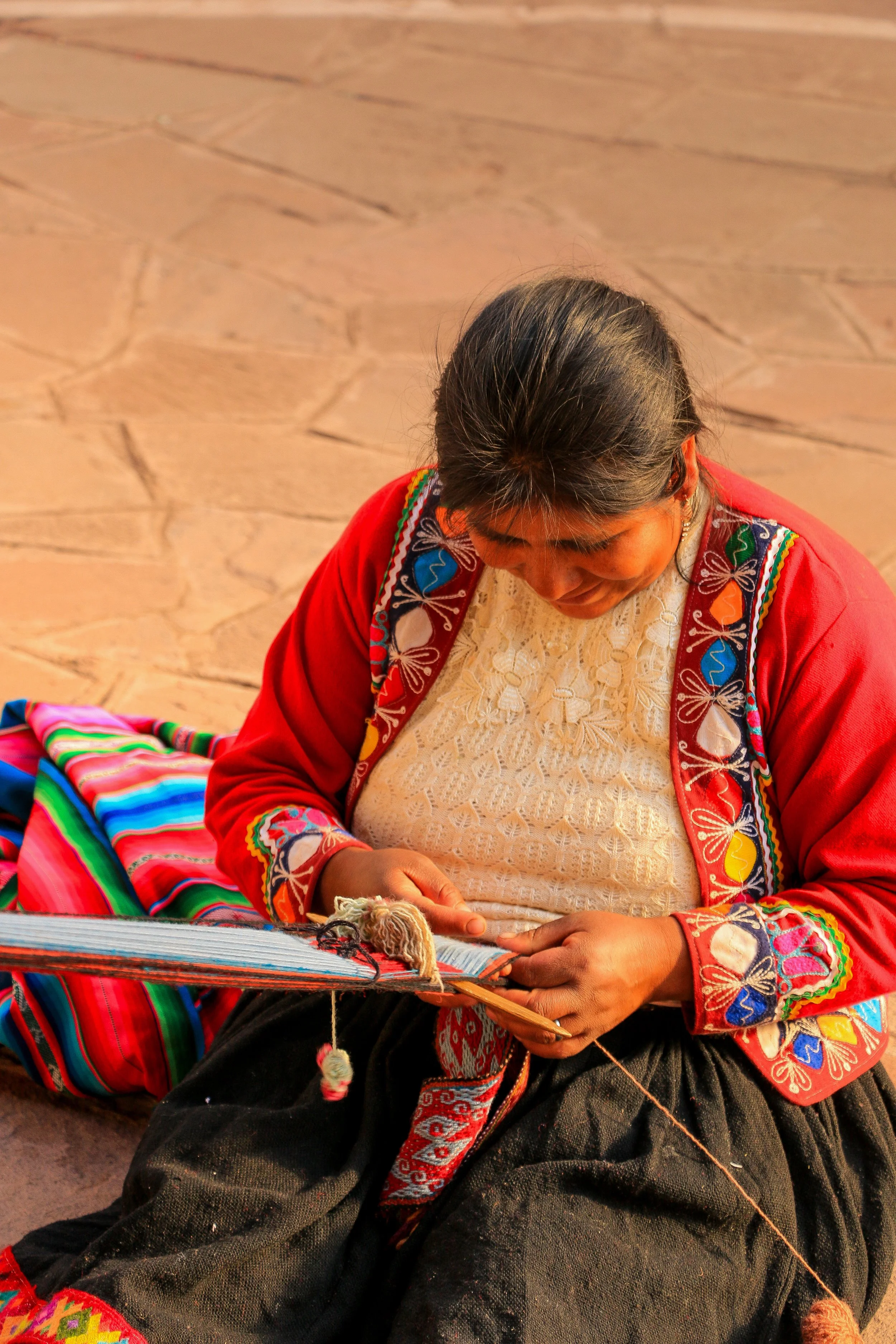 A woman sitting on the ground, wearing traditional embroidered clothing, weaving on a backstrap loom with colorful threads.