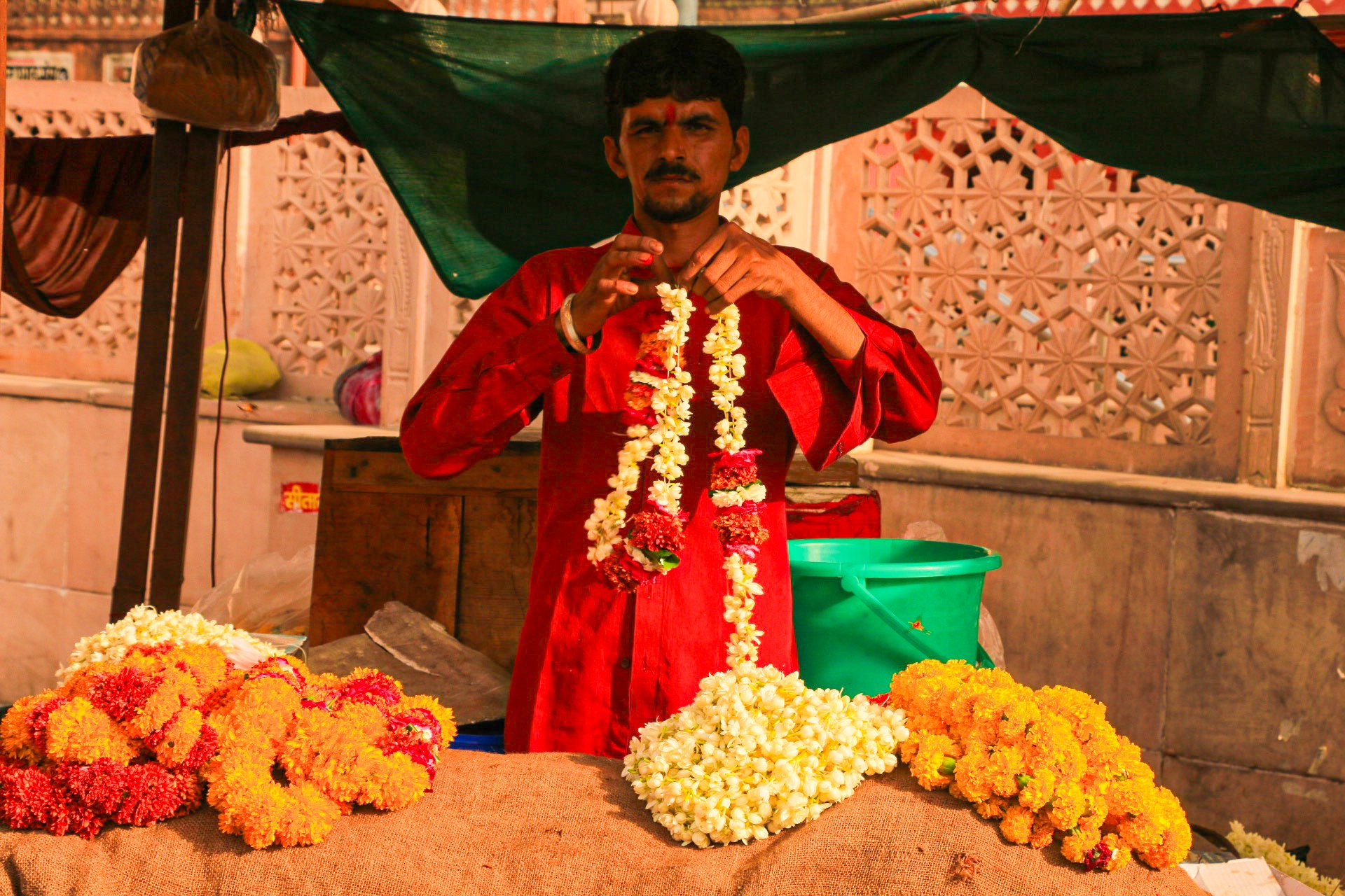 A man in traditional red attire arranging flower garlands at a flower stall with marigolds and jasmine flowers, under a green tent in an outdoor market.