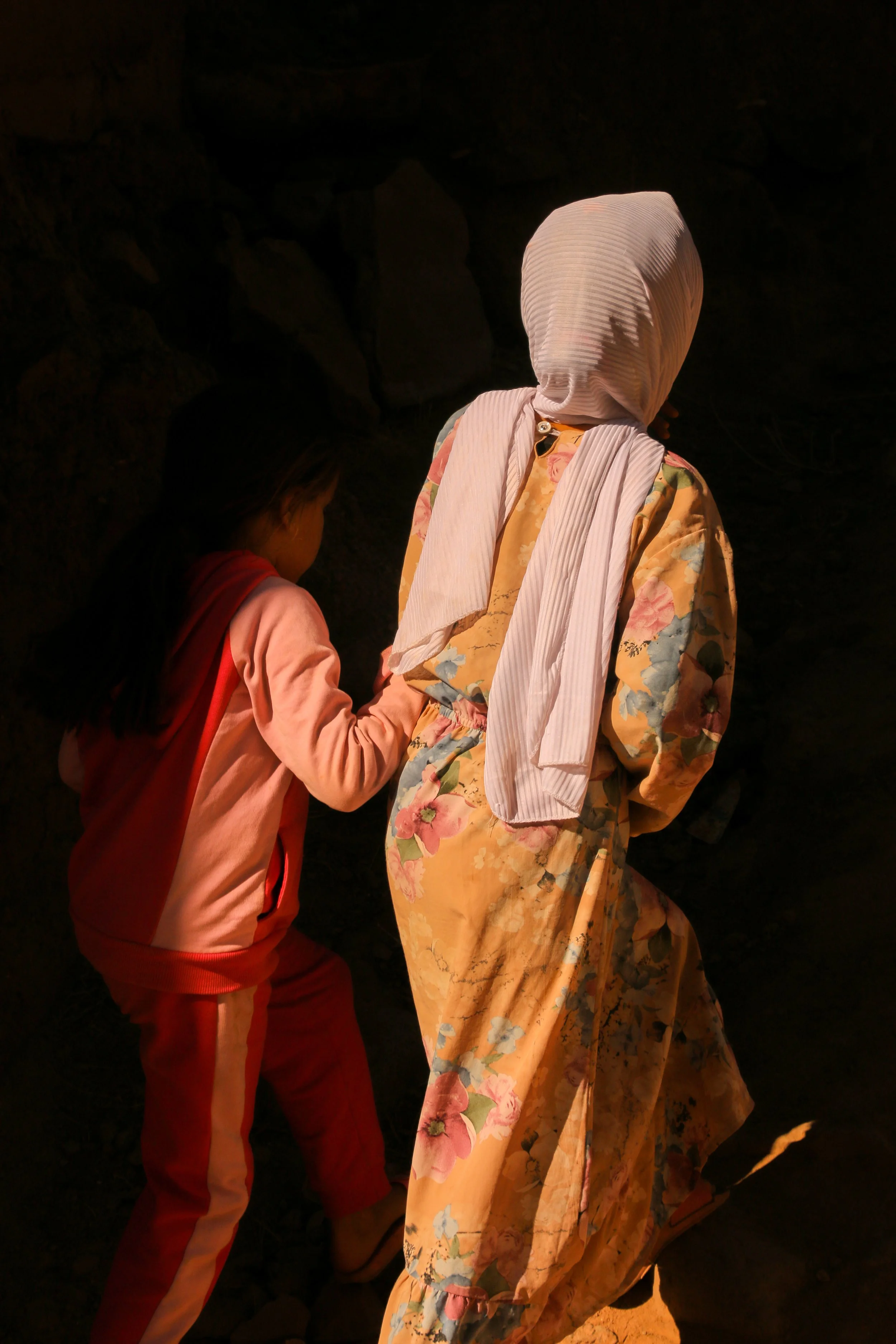 A woman dressed in a yellow floral dress with a white head covering holding hands with a young girl in pink clothing, in a dark setting with rocky background.
