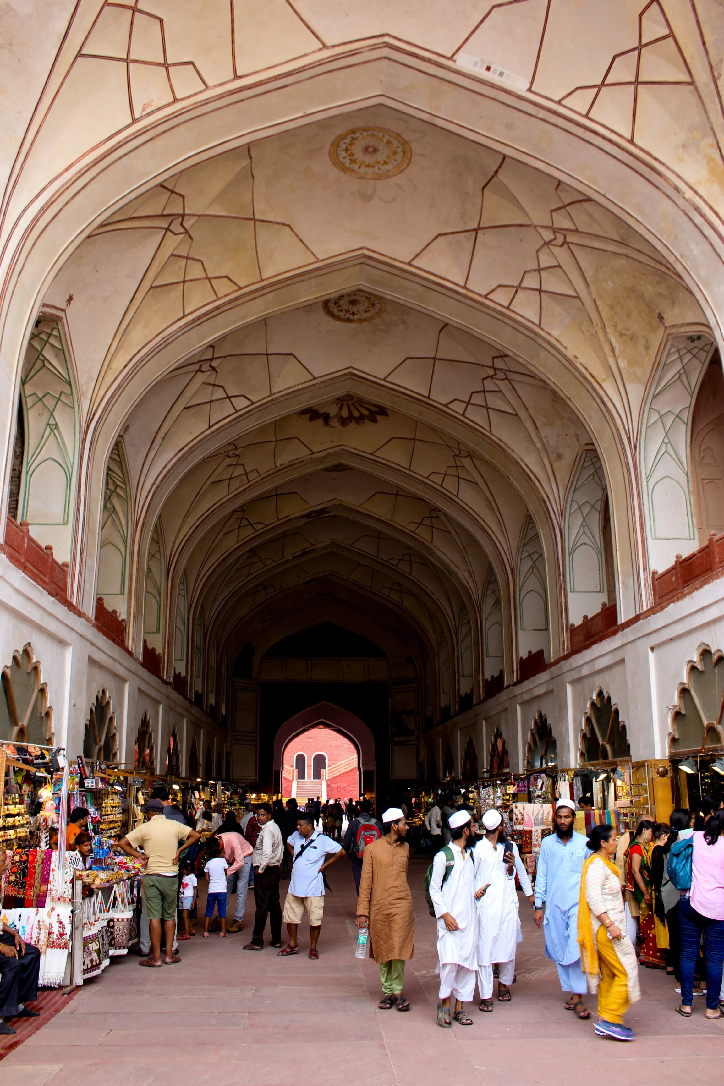 People shopping at outdoor market stalls under a grand, arched, and decorated ceiling in an open-air setting.