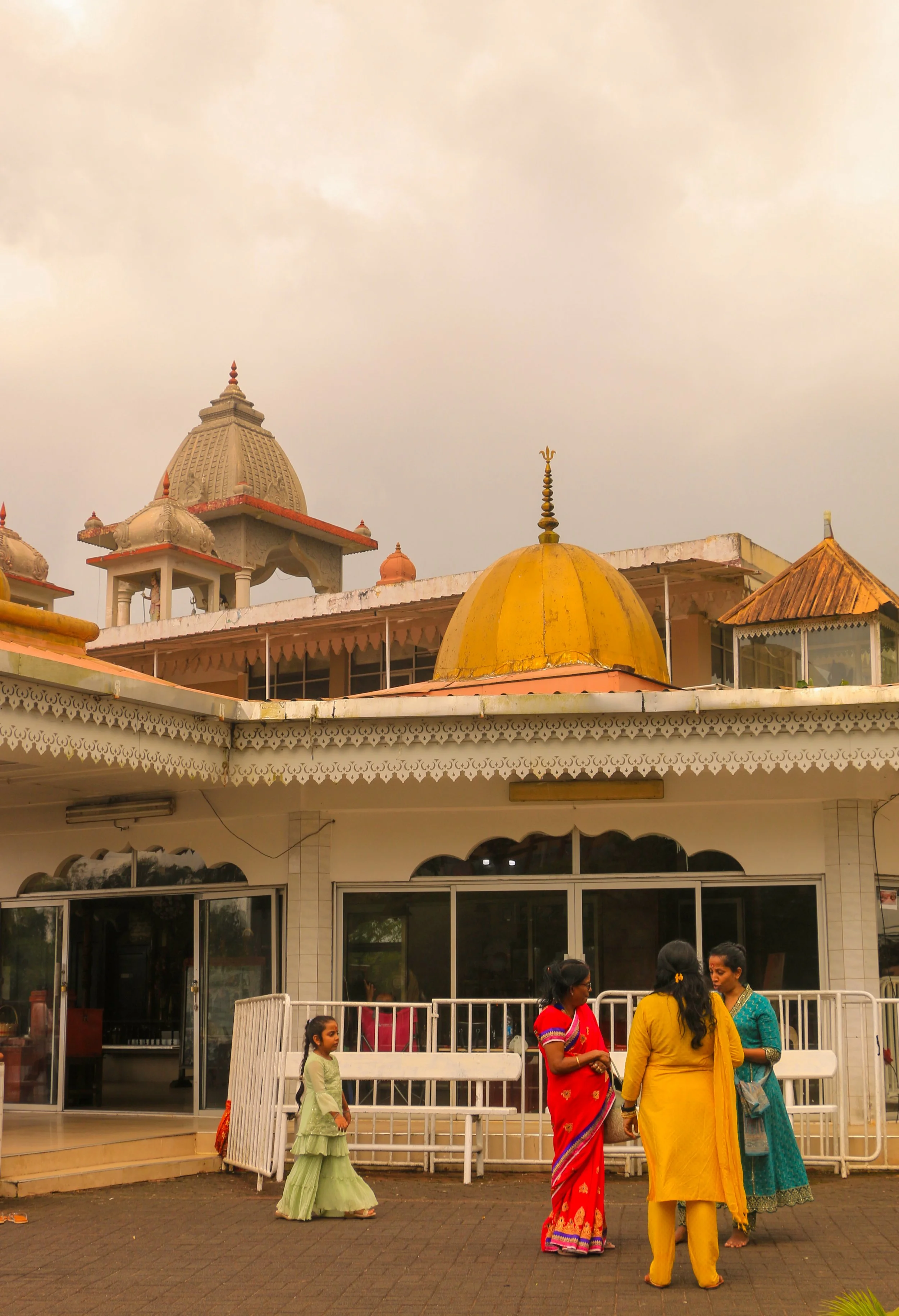 A group of women and a girl in colorful traditional Indian attire standing and conversing outside a temple with domed rooftops and intricate architecture.