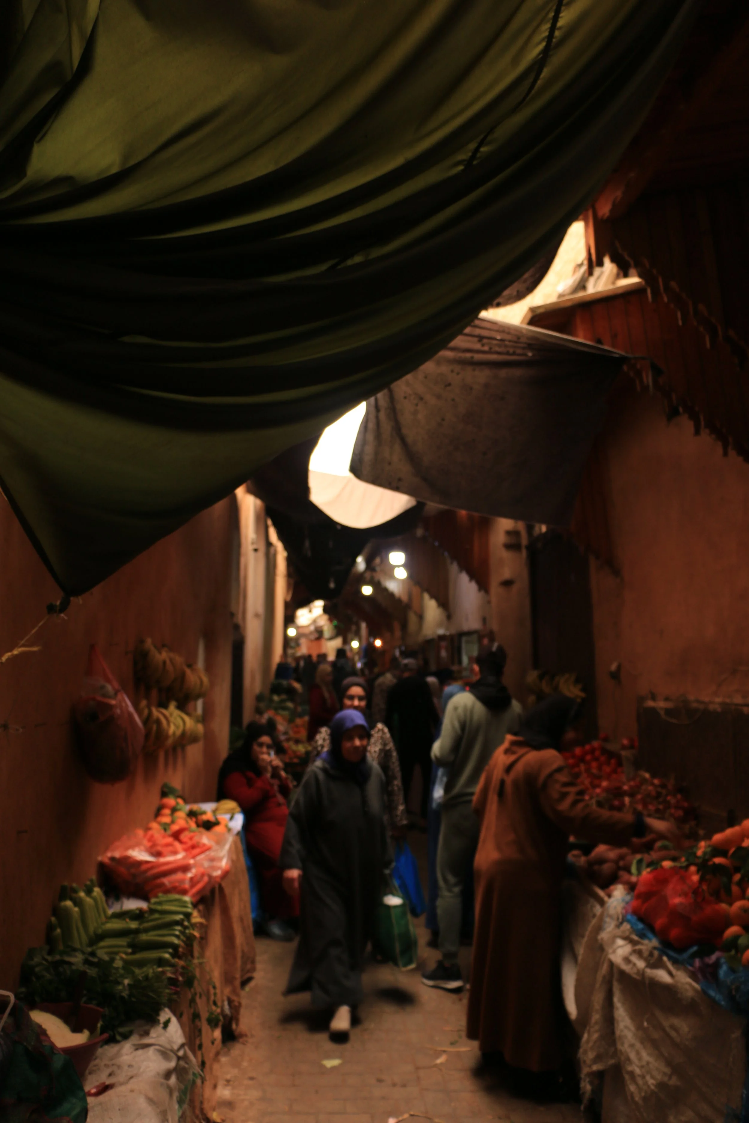 A bustling indoor market with vendors selling vegetables and fruits, and people shopping and walking through the narrow alleyway.