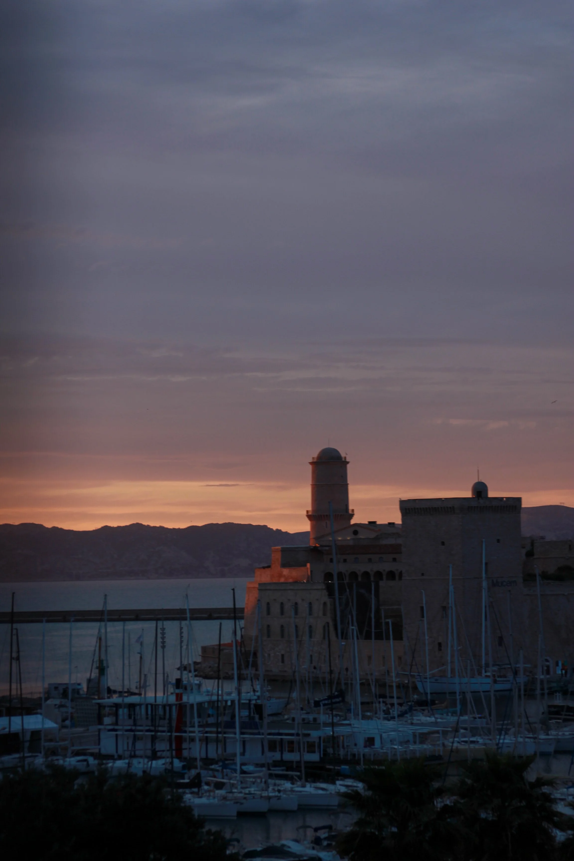 Sunset over a harbor with sailboats and historic buildings, mountains in the background.