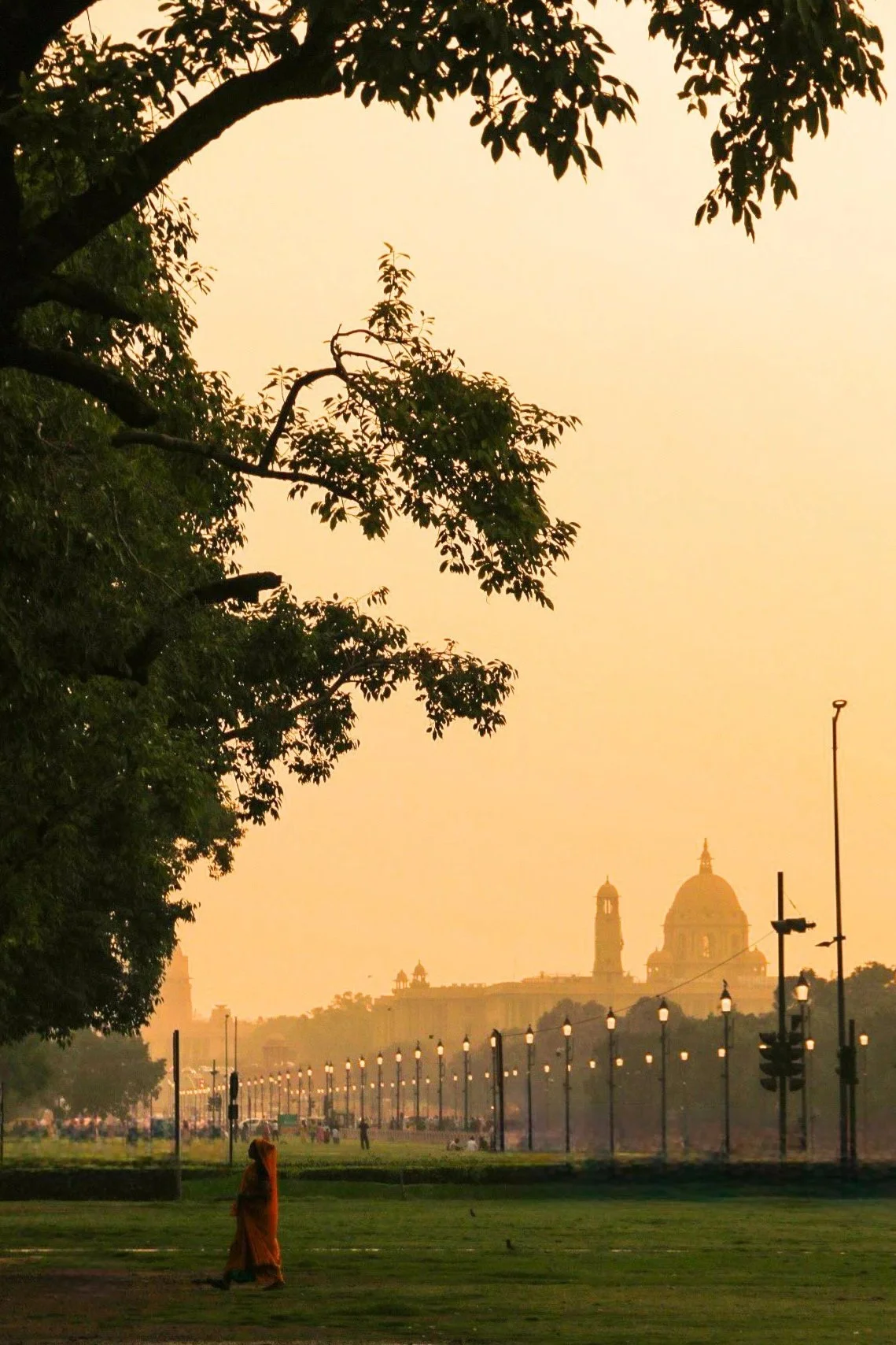 A city park at sunset with trees and a distant view of a historic building with domes and towers, and a person dressed in orange walking on the grass.