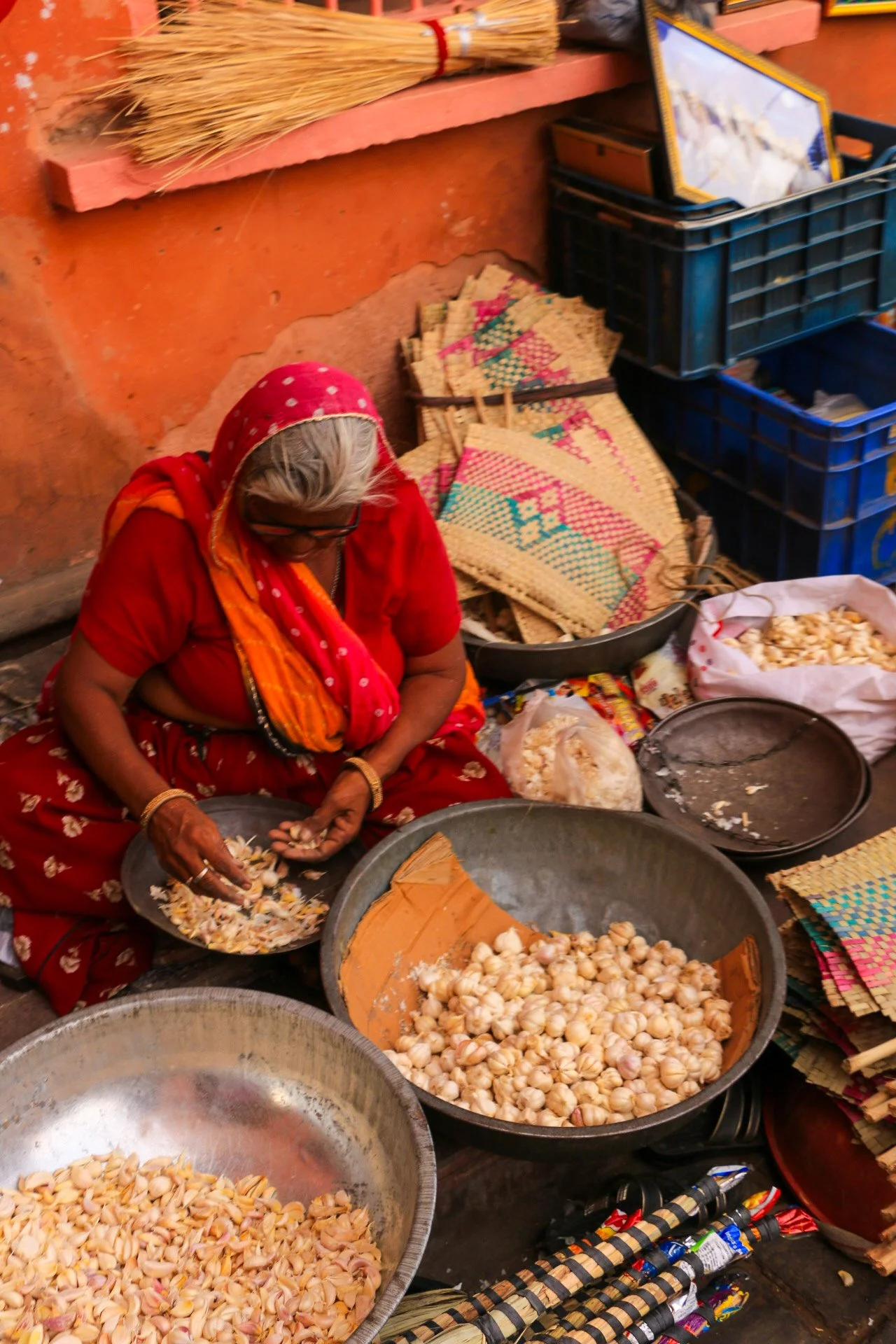 An elderly woman in traditional Indian attire, including a red sari with gold floral patterns and a red headscarf, is sitting on the ground peeling garlic. She is surrounded by large bowls filled with garlic cloves and garlic peels, with woven mats and other decorative items nearby.