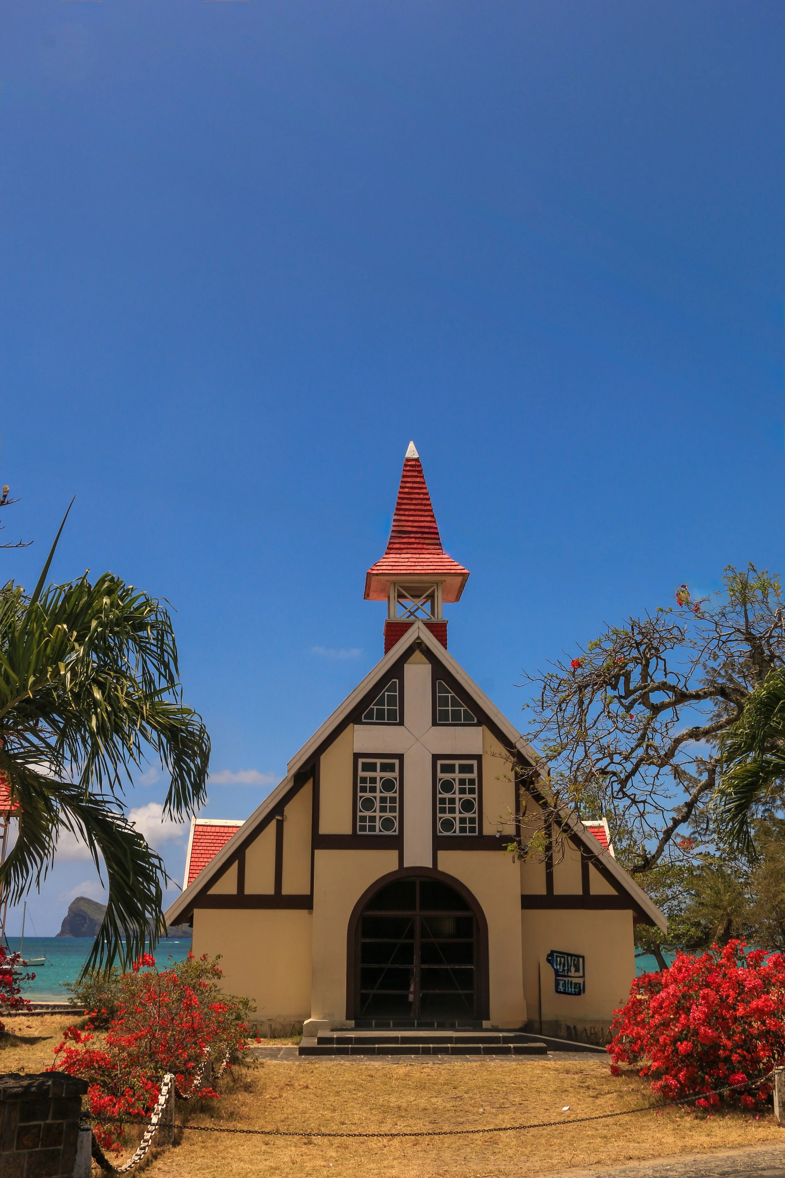 A small, Tudor-style building with a steeply pitched roof and a tall red spire, situated in a tropical setting with palm trees, flowering shrubs, and a view of the beach and water in the background.