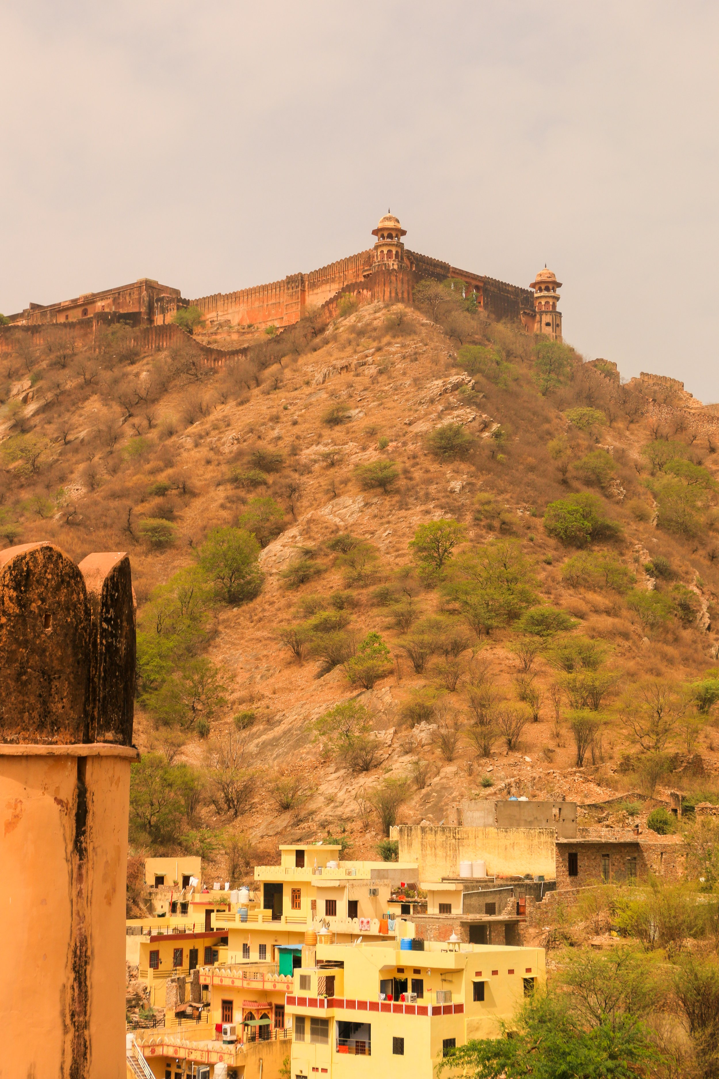 A hilltop fort with watchtowers and walls overlooking a small residential area, with dry, rocky terrain and sparse green trees.