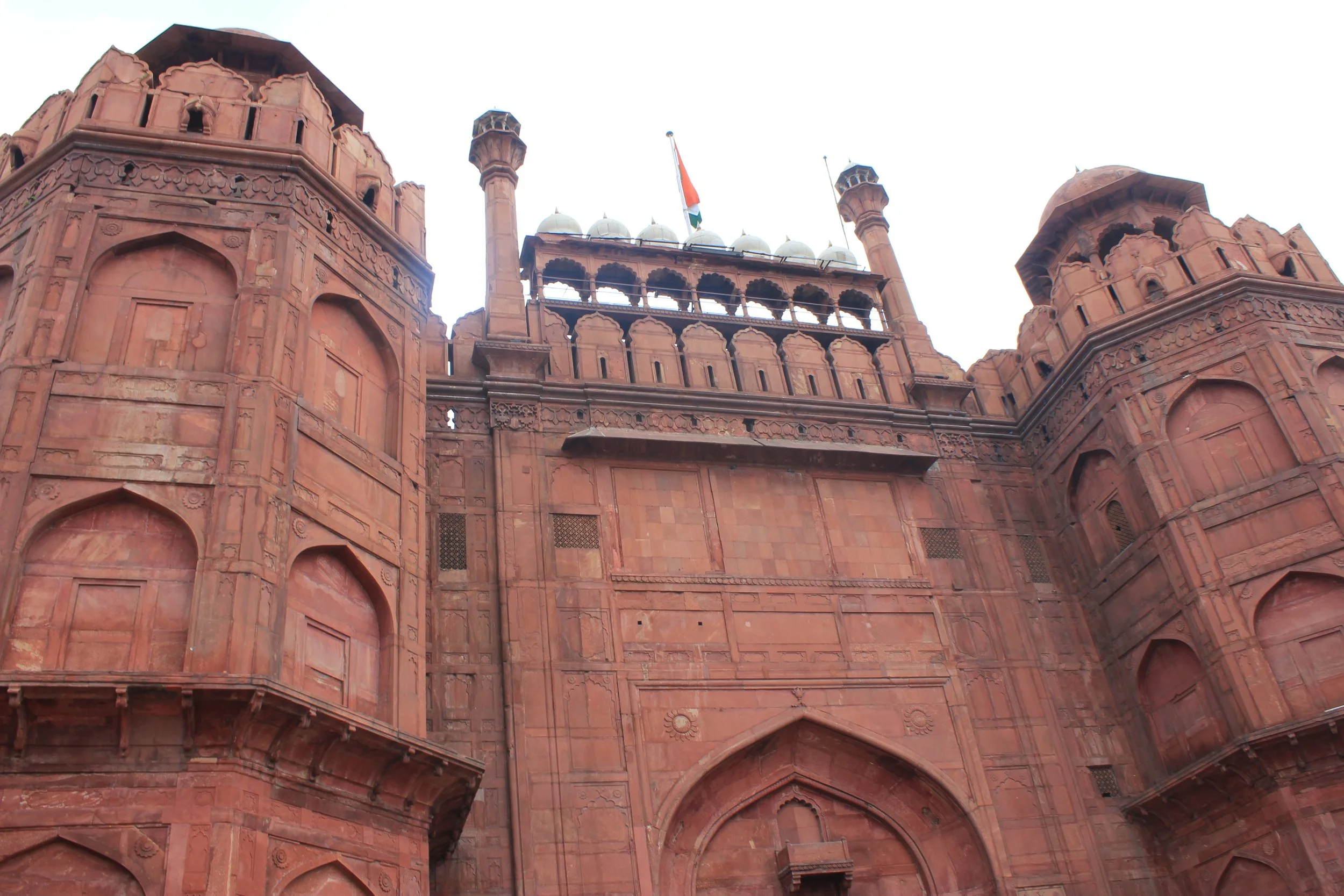 The facade of a historic red sandstone building with intricate architectural details and multiple arched windows, topped with small domes and minarets, with an Indian flag flying at the top.