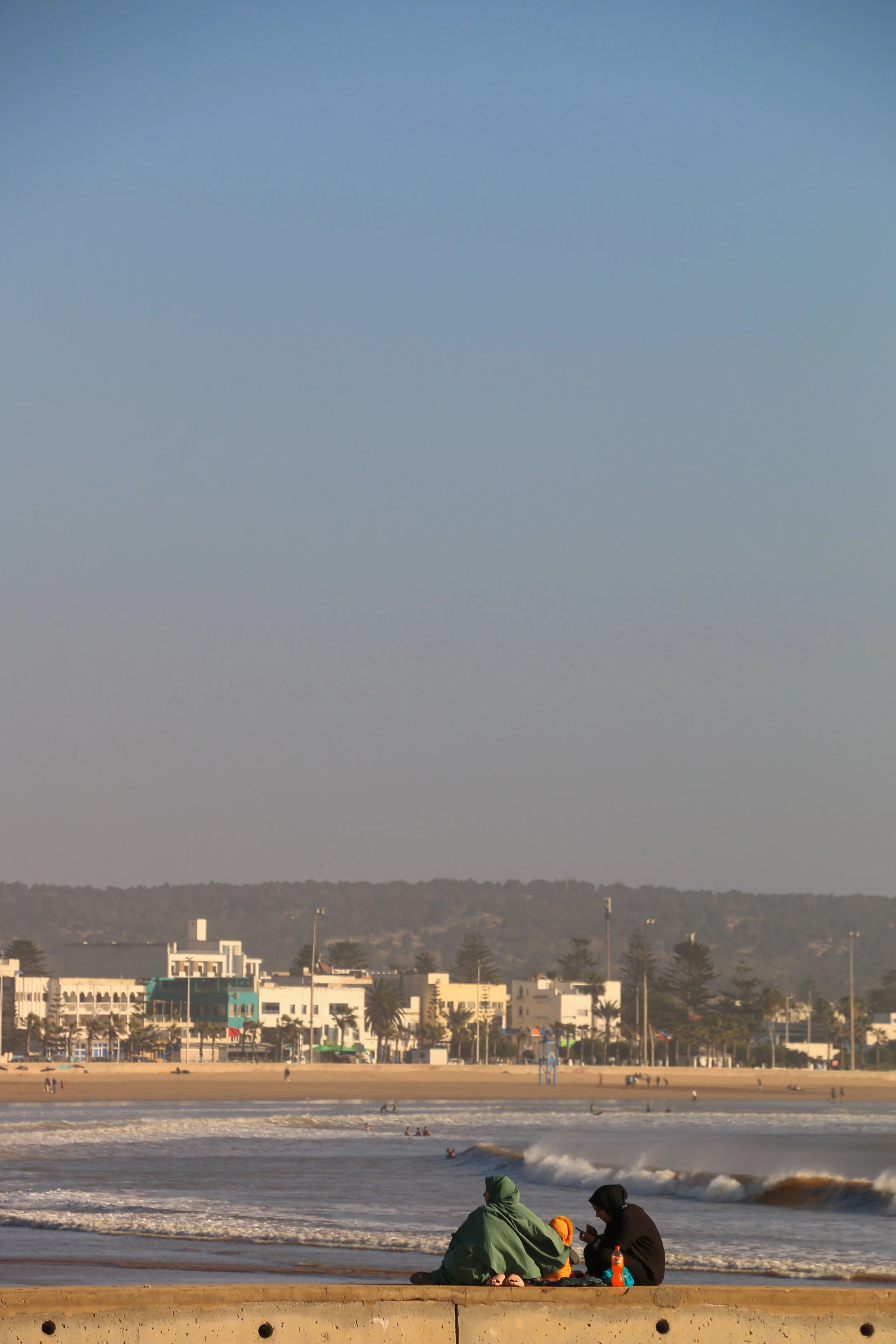People sitting on a beach at sunset, with waves crashing onto the shore and a cityscape in the background.