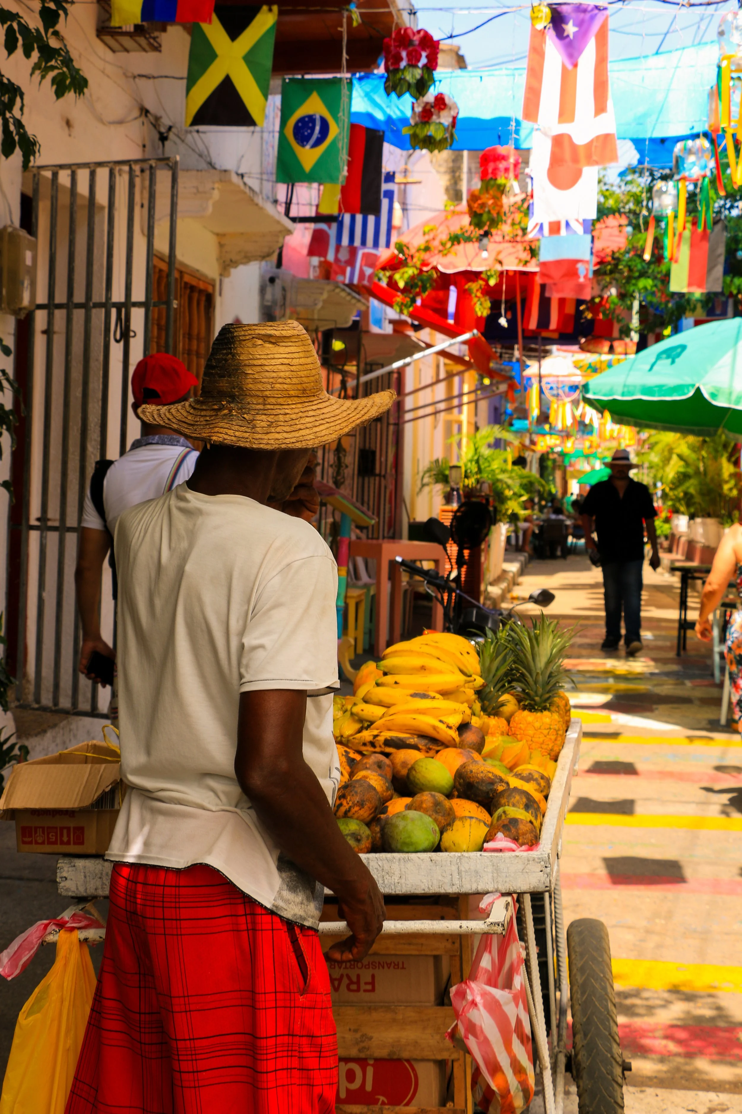 A street market scene in a tropical location with a vendor selling bananas, pineapples, and papayas. Colorful flags, including Jamaican, Brazilian, and American flags, hang overhead. People walk along the street under umbrellas and bright sunlight.