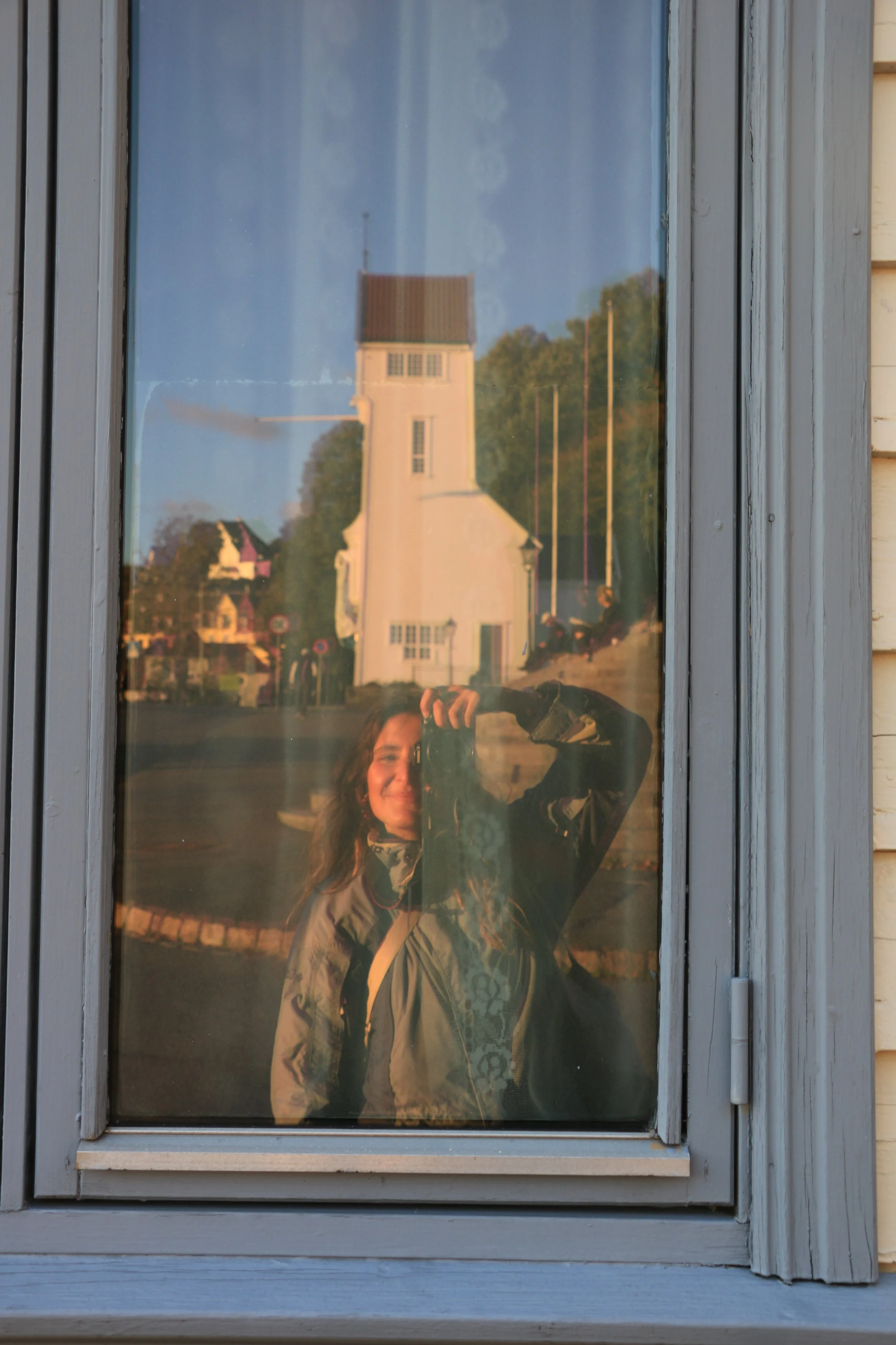 Reflection of a woman with curly hair taking a photo with a camera in a window, with a white church and trees in the background during sunset.