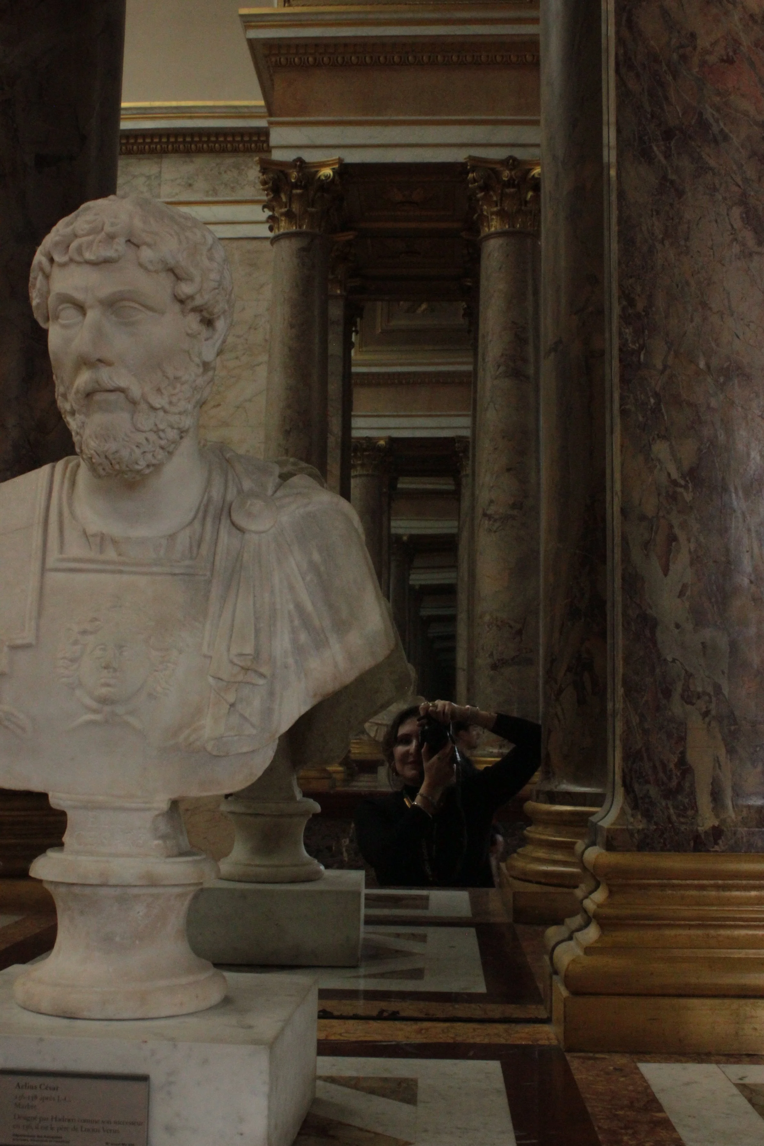 Reflection of a woman taking a photo in a mirror beside a marble bust sculpture of a bearded man in an ornate hall with marble columns and detailed architecture.
