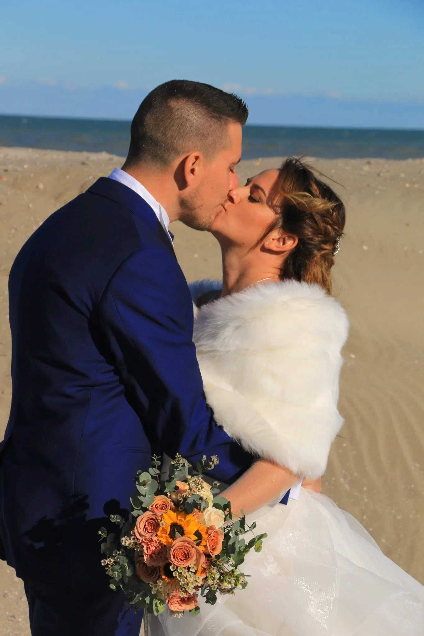 A couple dressed in wedding attire sharing a kiss on a sandy beach with the ocean and blue sky in the background.