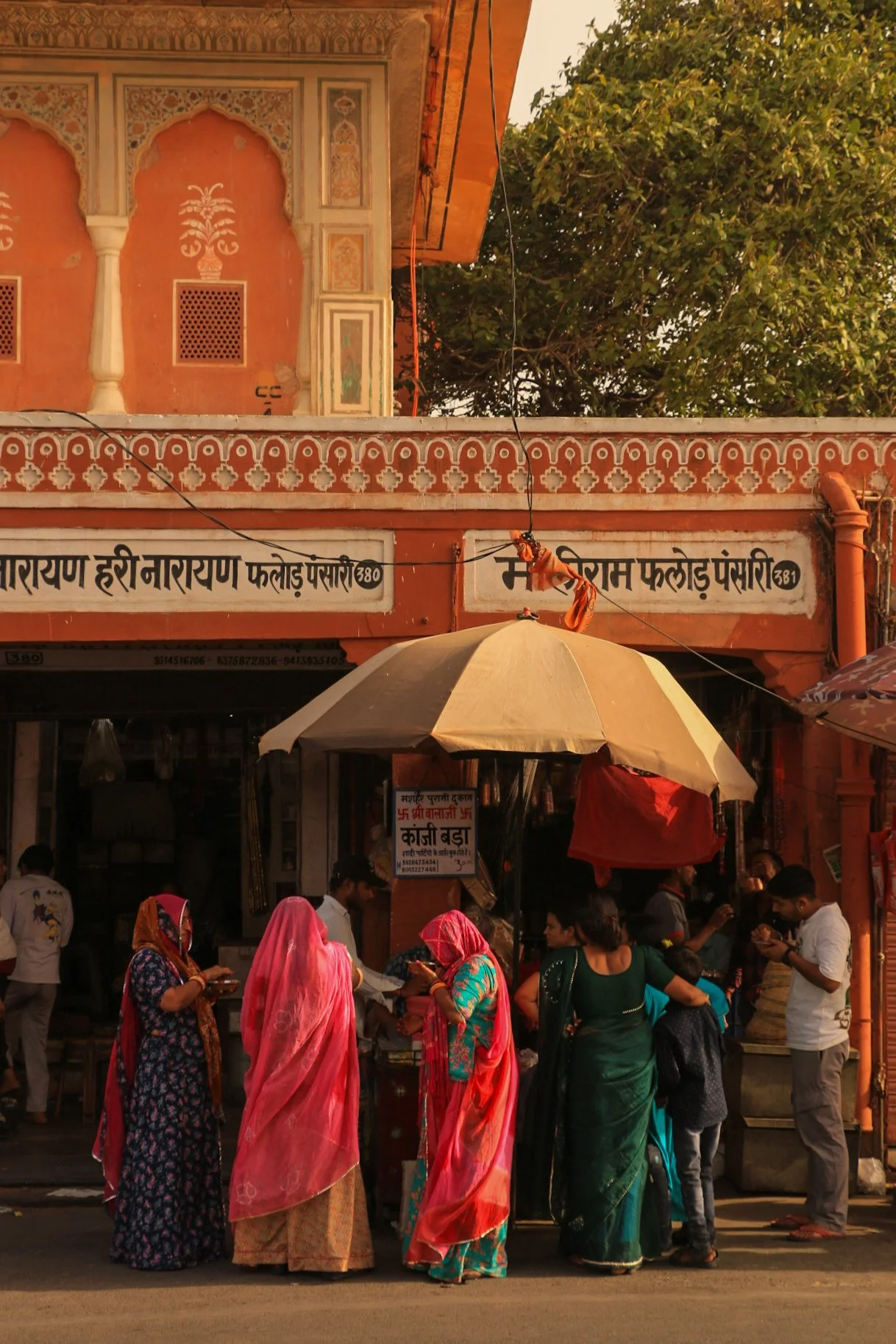 People gathered under a large yellow and white umbrella outside a colorful Indian building, some wearing traditional sarees, and others in modern attire, with trees in the background.