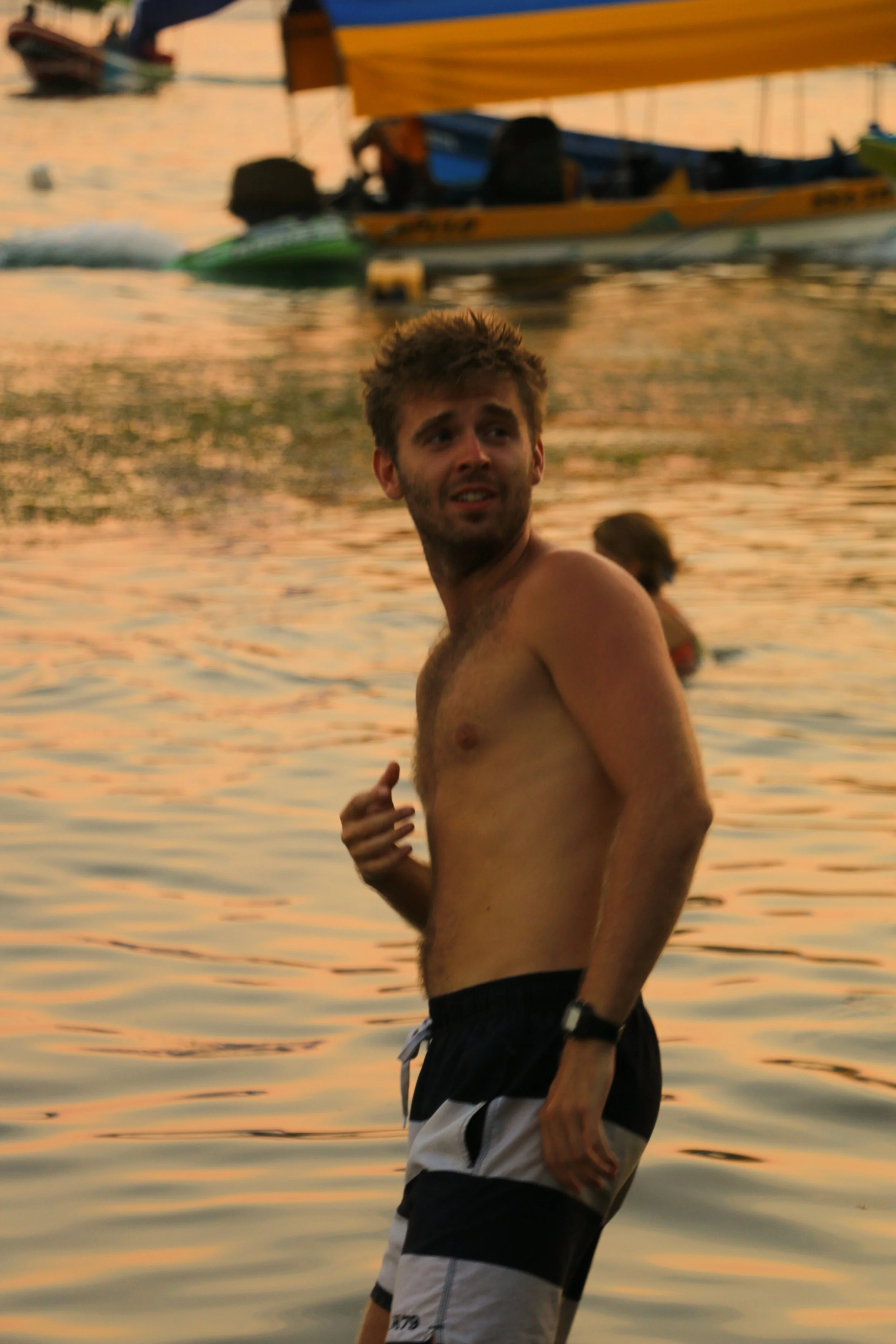 A young man with light brown hair and facial hair, shirtless, wearing black and white swim trunks, standing in water during sunset, with boats in the background.