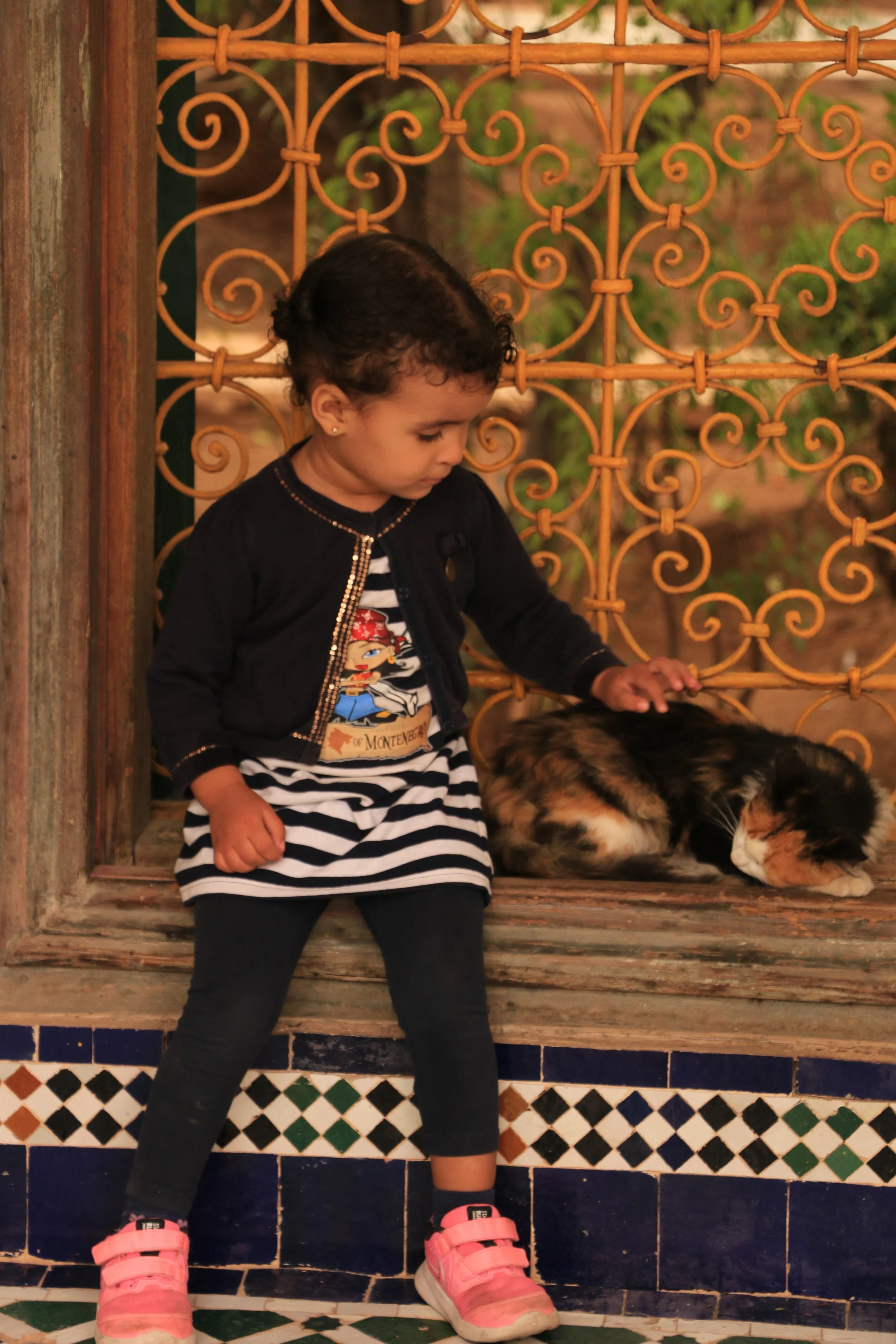 A young girl with short dark hair, wearing a black jacket, striped dress, black leggings, and pink shoes, is petting a calico cat that is lying on a wooden windowsill behind an ornate orange metal fence.