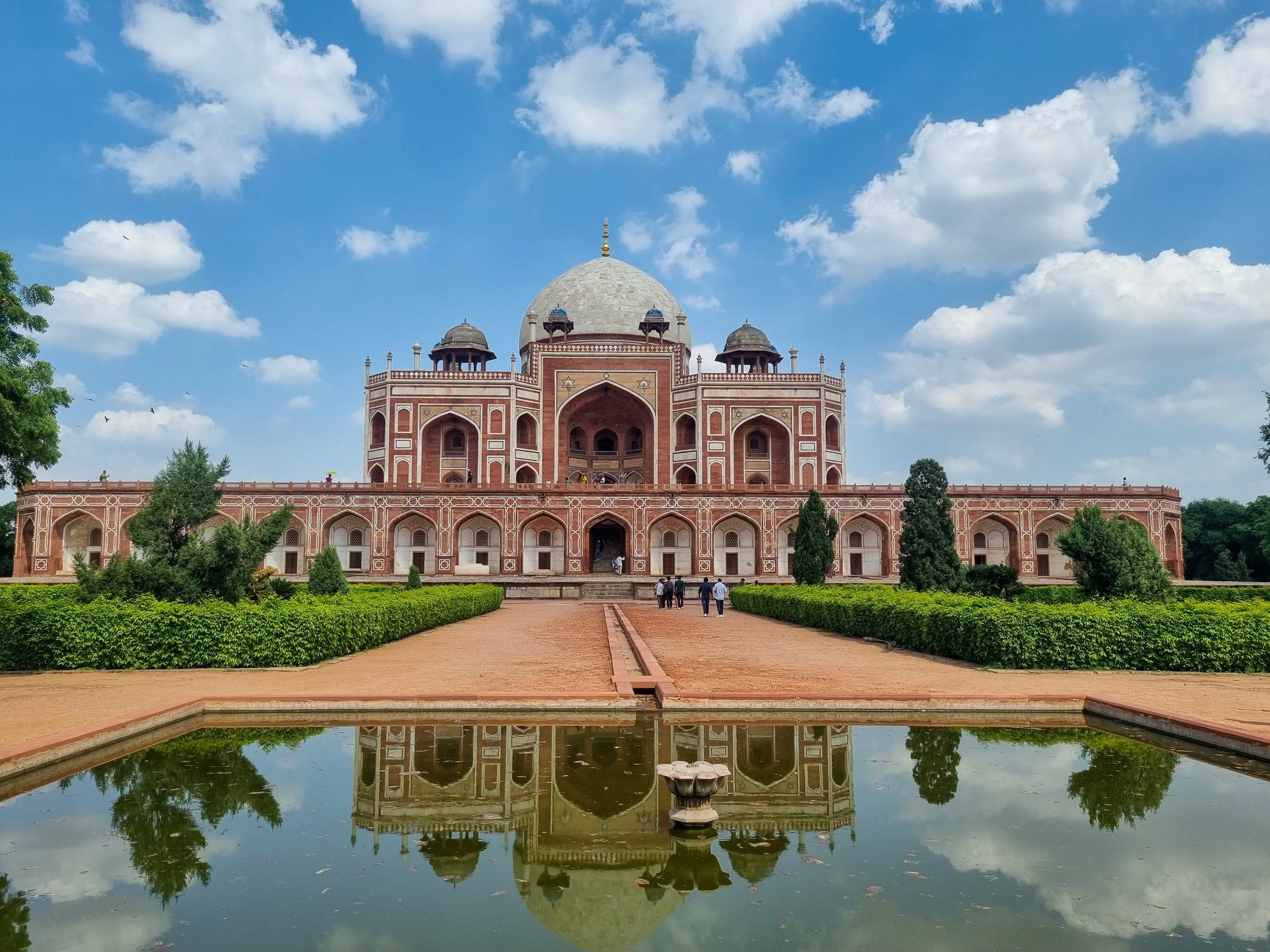 The Taj Mahal, a white marble mausoleum, with a large central dome and smaller domes on either side, surrounded by lush greenery and a reflecting pool in the foreground, under a partly cloudy sky.