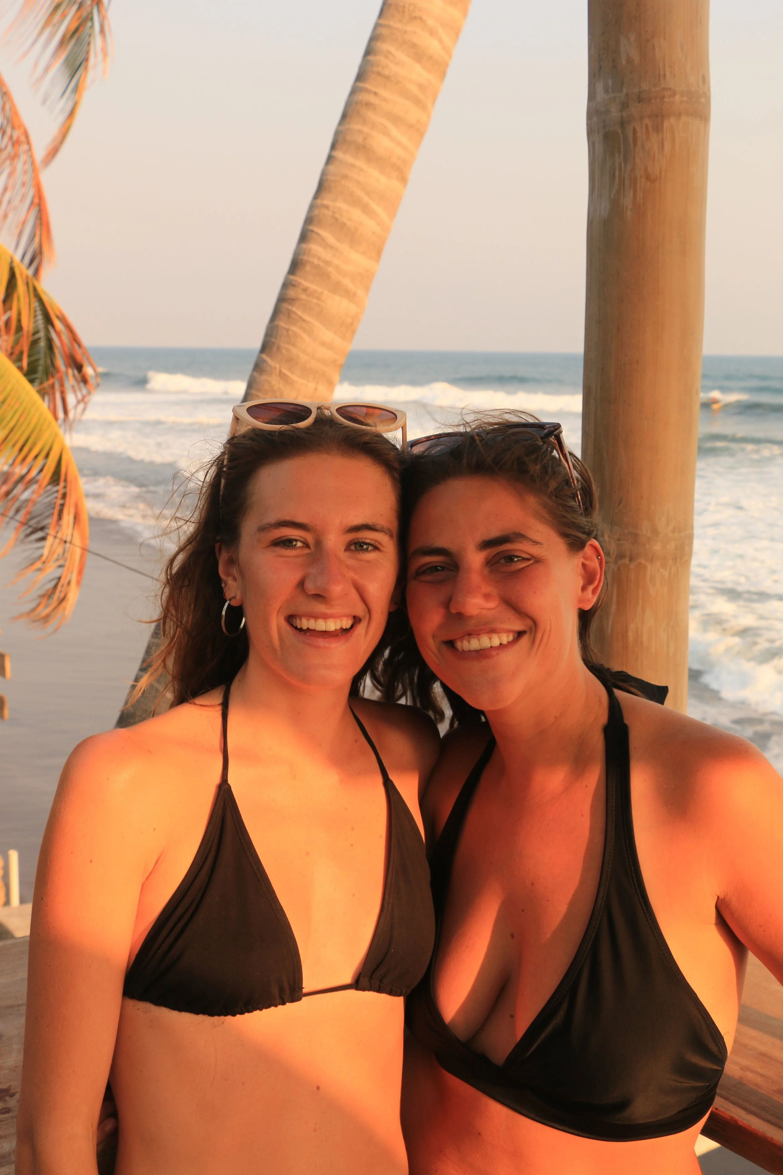 Two women in black bikinis smiling at the beach with ocean waves and palm trees in the background during sunset.