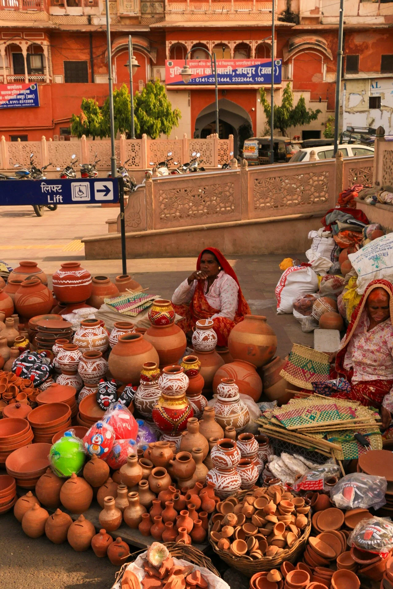 Street market with clay pottery on display, two women selling pottery, in front of pink historic building, with parked motorcycles and a sign for a lift and accessible parking nearby.