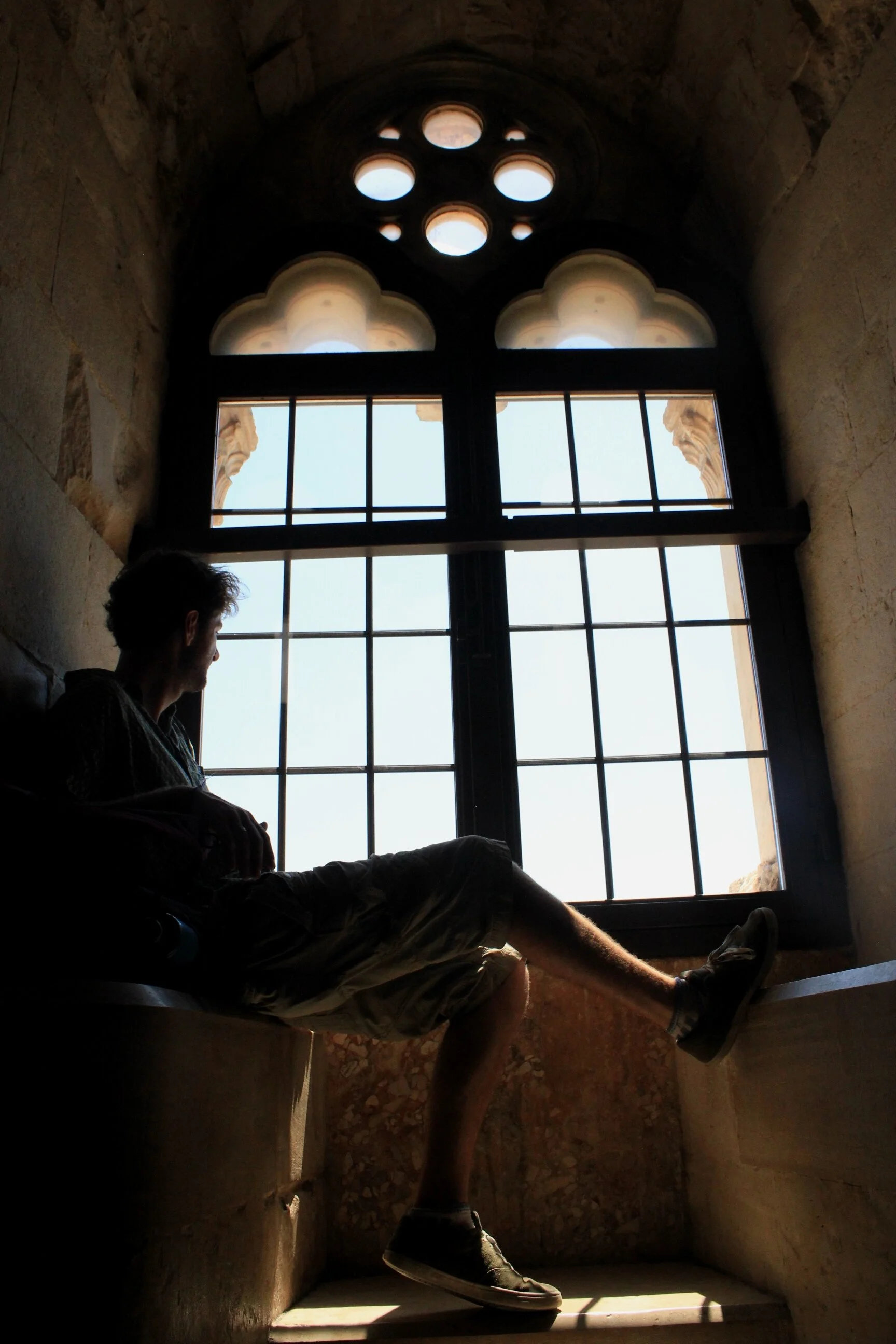 A person sitting and looking out a large, decorative window inside a stone building.