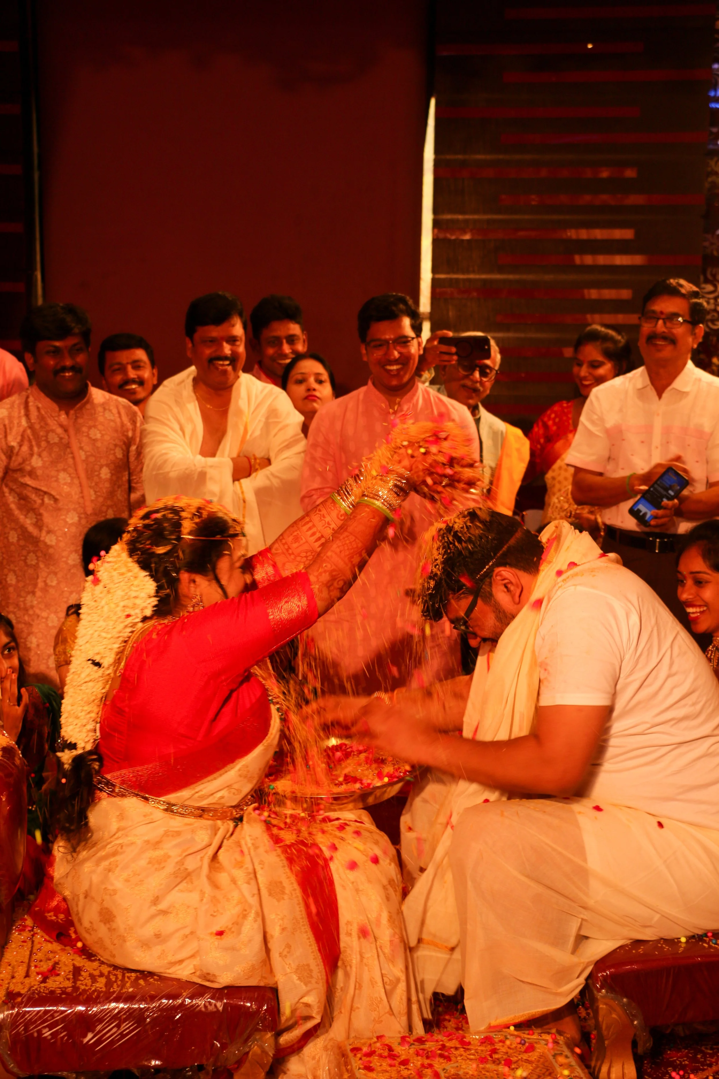 A traditional Indian wedding ceremony with a bride and groom performing a ritual, surrounded by family and friends, with flower petals and colorful attire.