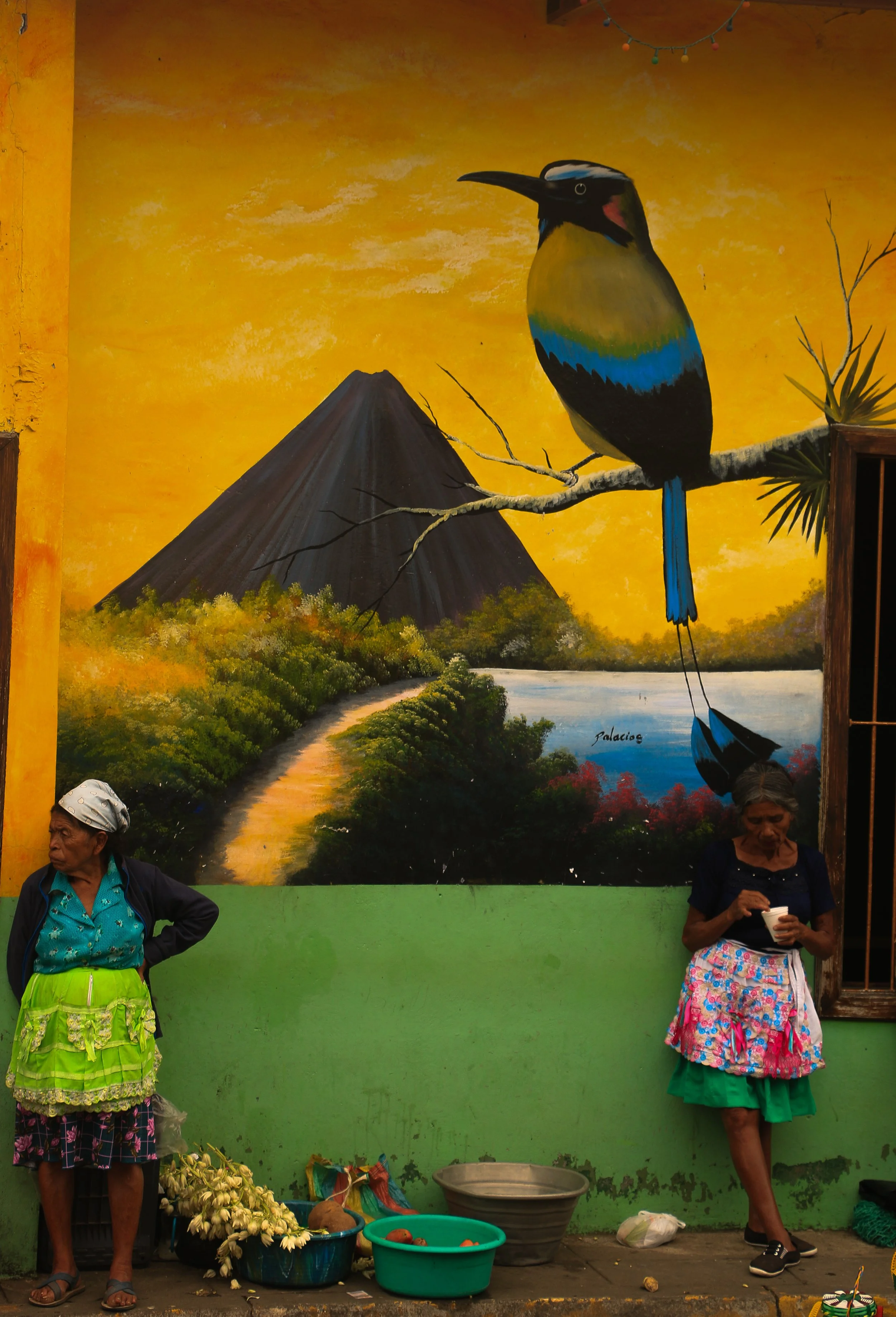 A mural of a colorful bird perched on a branch in front of a mountain and river landscape, with two women standing nearby outdoors.