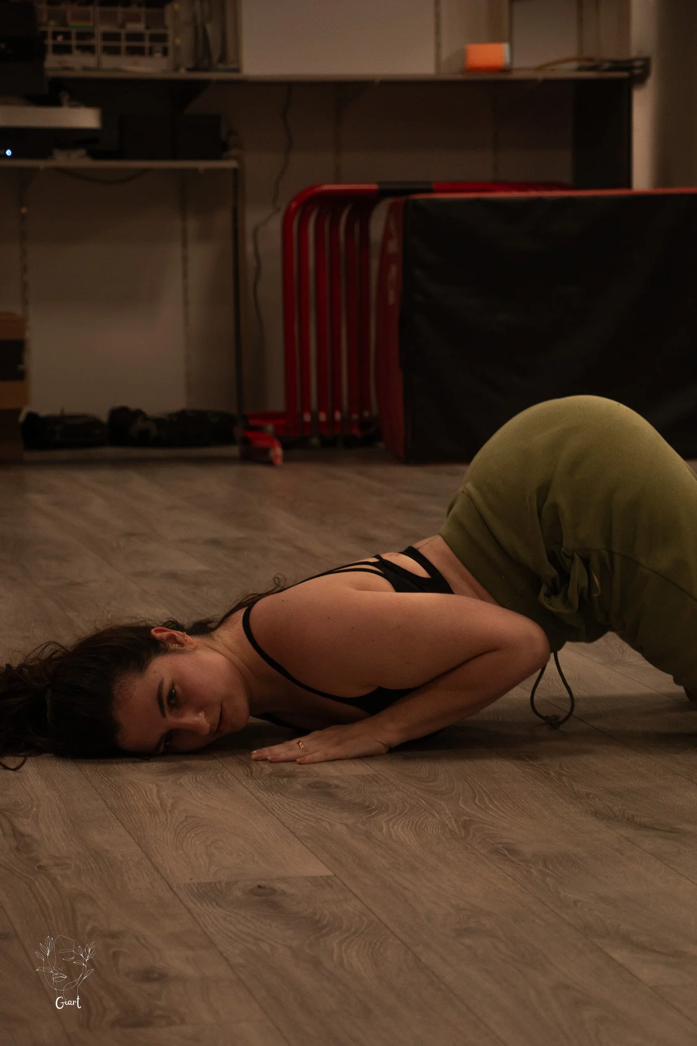 A woman doing a floor exercise pose on a wooden floor, with her head tilted to the side and her body in a low crawl position.
