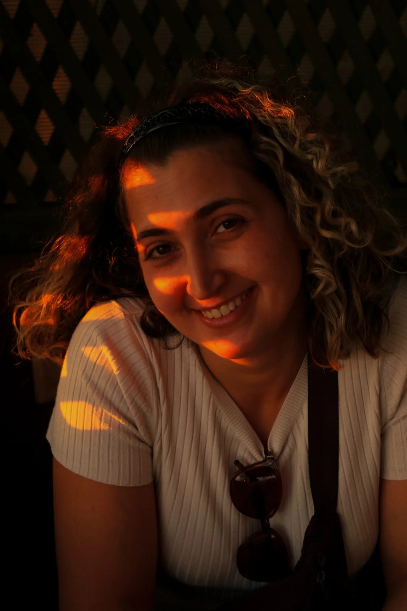 A woman with curly hair and sunglasses hanging from her shirt smiling in warm sunlight.