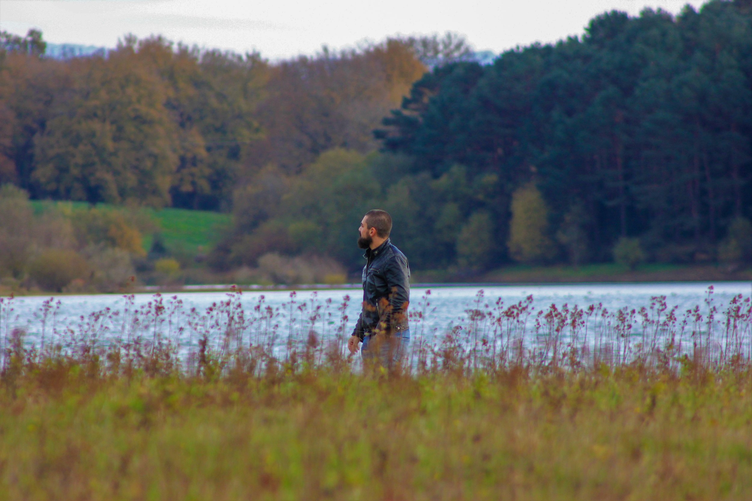 A man standing in a field near a lake with trees in the background on a cloudy day.