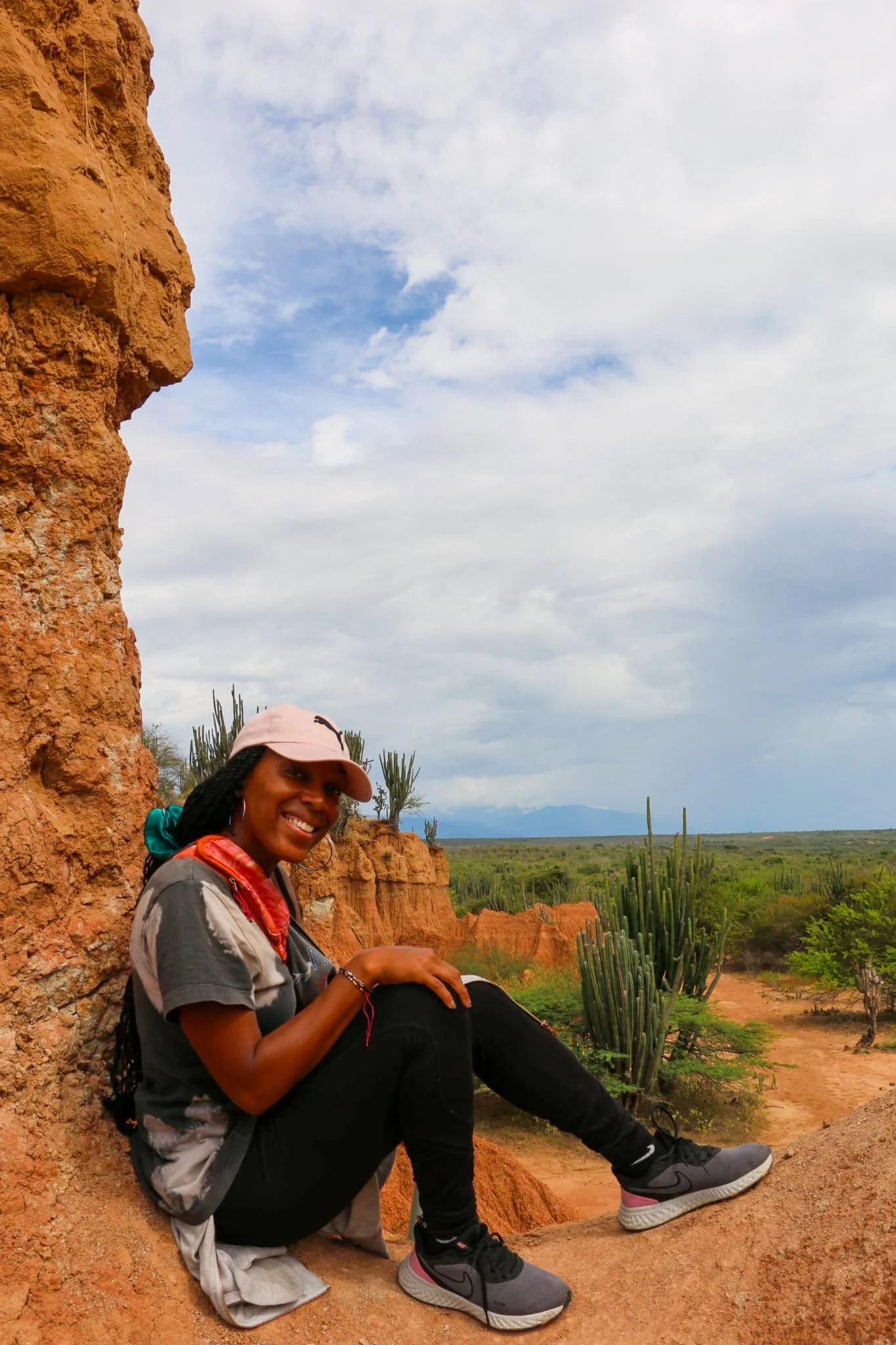 A woman with dreadlocks wearing a pink cap, black pants, gray sneakers, and a bandana around her neck, sitting on a rock formation in a desert landscape with cacti and orange cliffs, smiling at the camera.