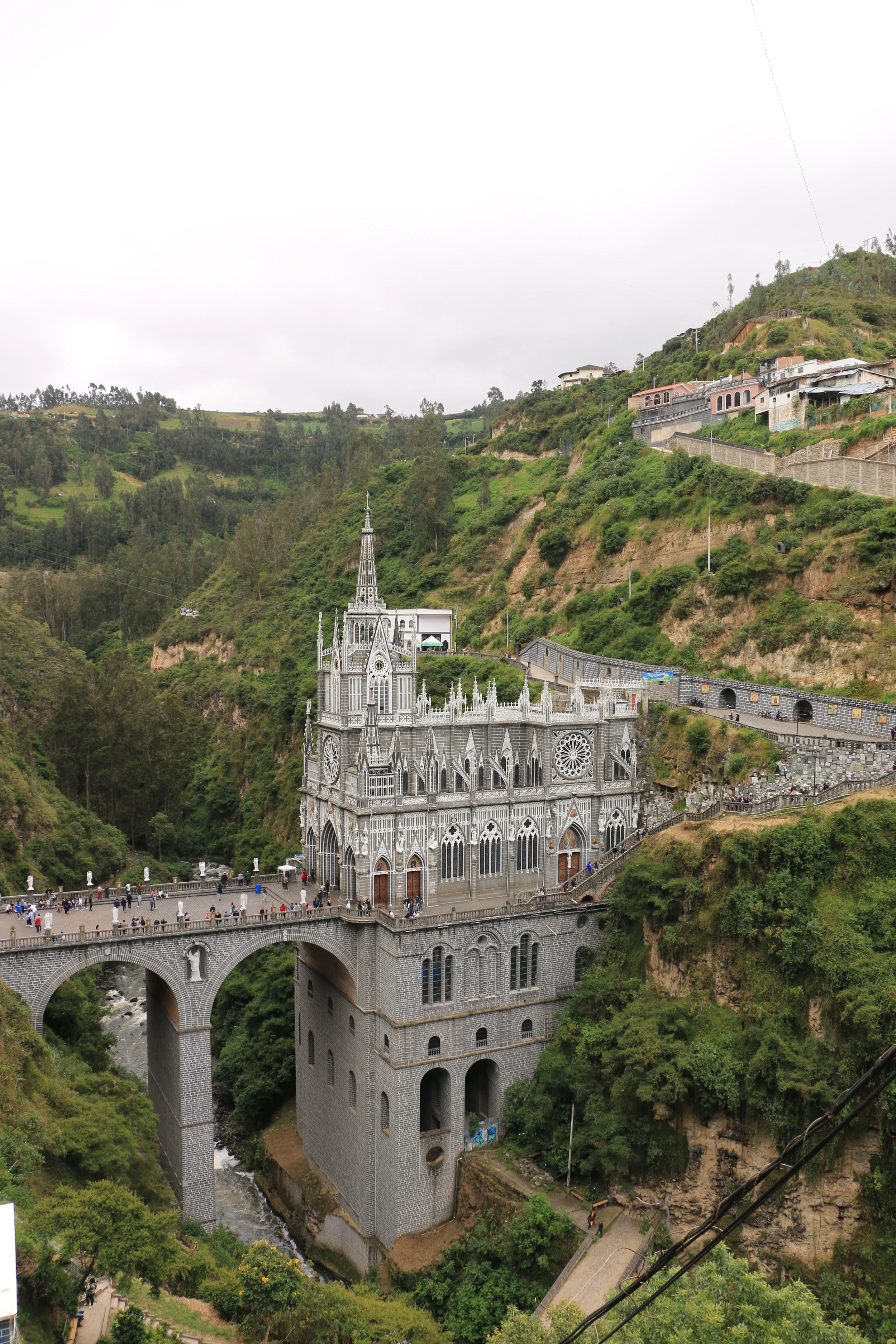 A church built on a hill with a bridge leading to its entrance, surrounded by green vegetation and houses on the hillside.
