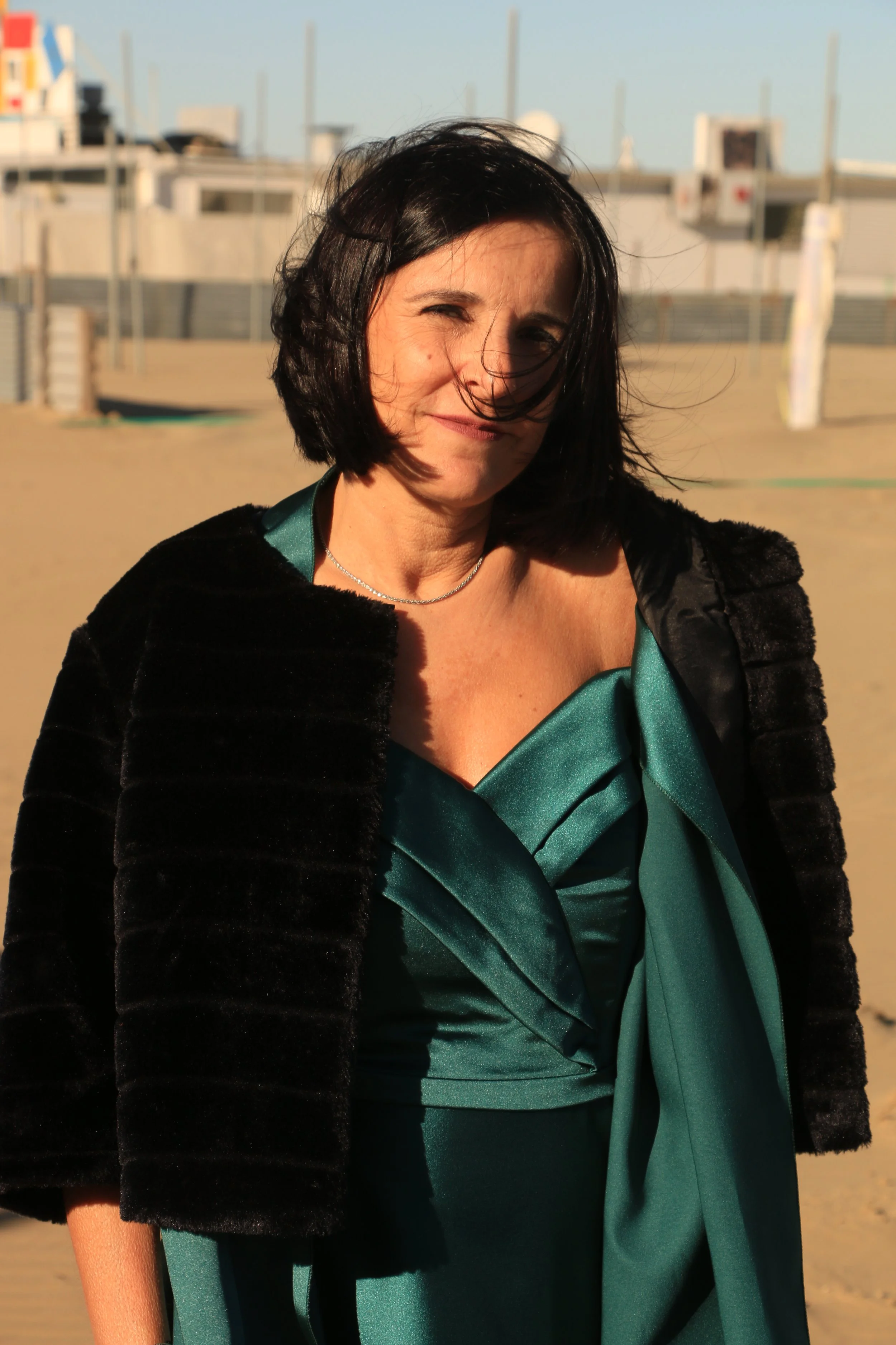 A woman with short dark hair wearing a teal dress and black jacket standing on a sunny beach.