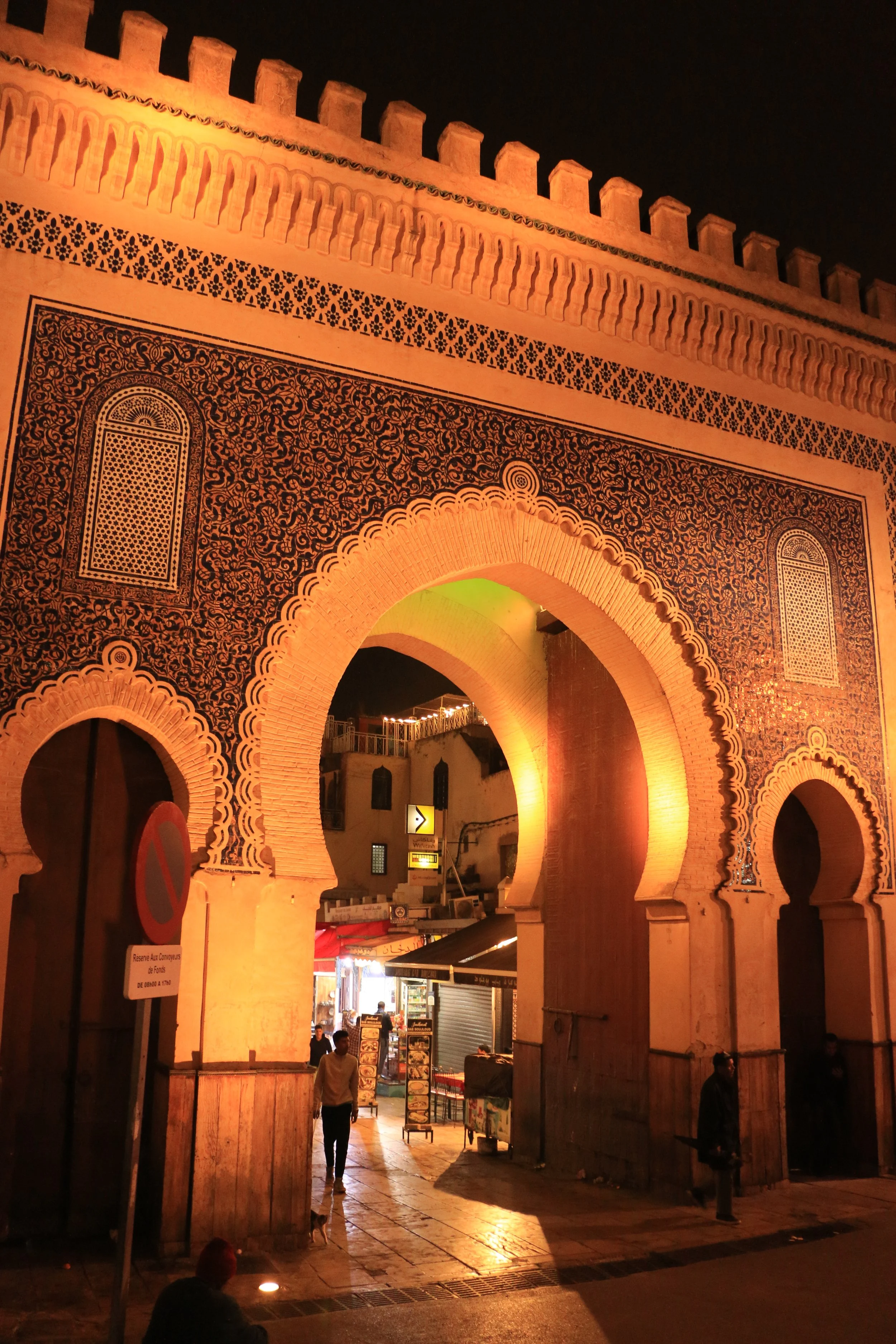 Night view of an illuminated traditional Moroccan city gate with intricate tile work and arches, with people and small shops in the background.
